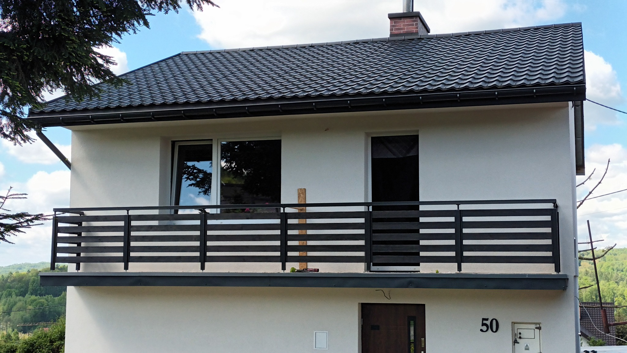 Nowoczesny dom z białymi ścianami i czarnym dachem. Na pierwszym planie balkon z czarną, poziomą balustradą. Widoczny numer domu '50'.
