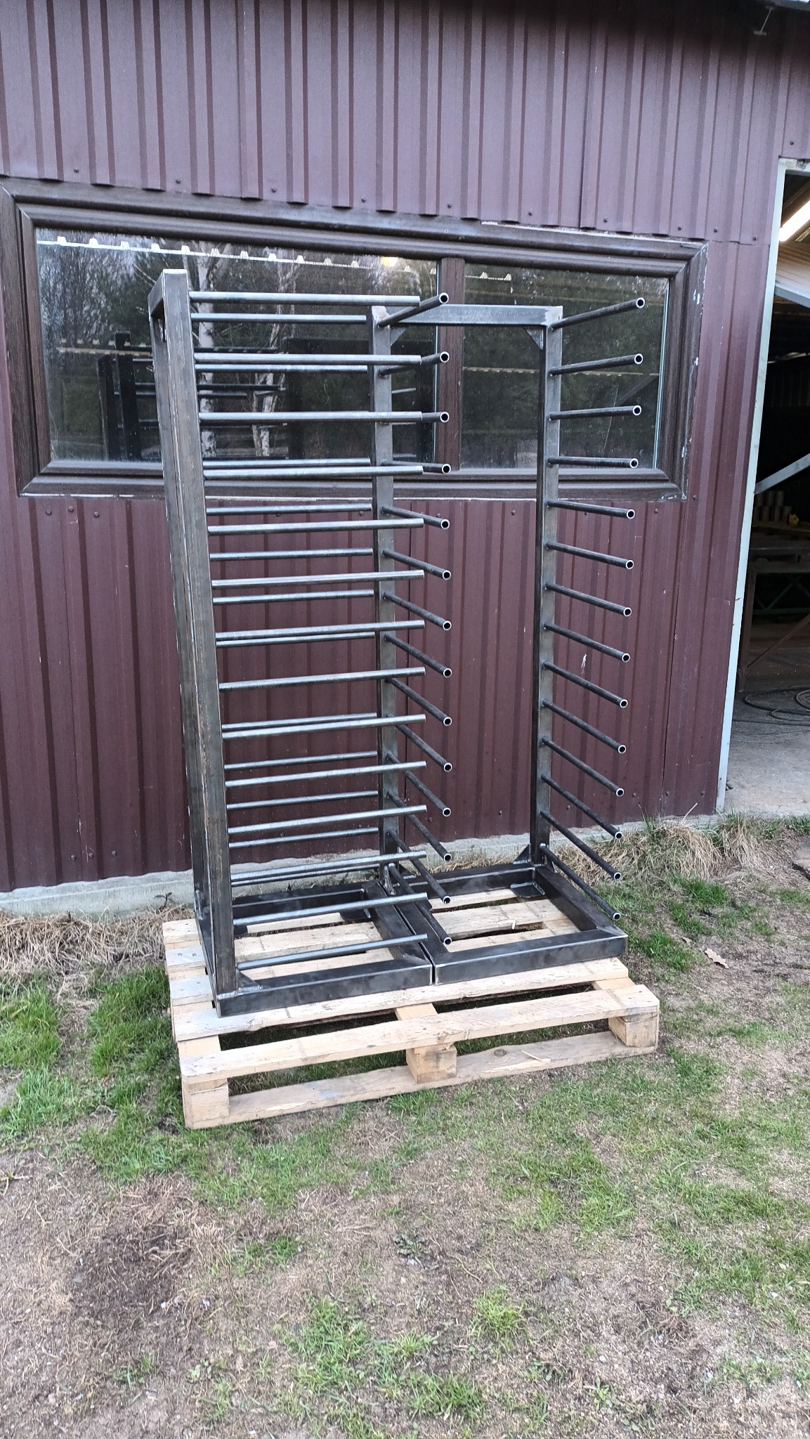 Metalowa konstrukcja regału na palecie, na zewnątrz budynku z brązową elewacją. Widoczne rzędy rurek i spawane elementy ramy. Konstrukcja do przechowywania.