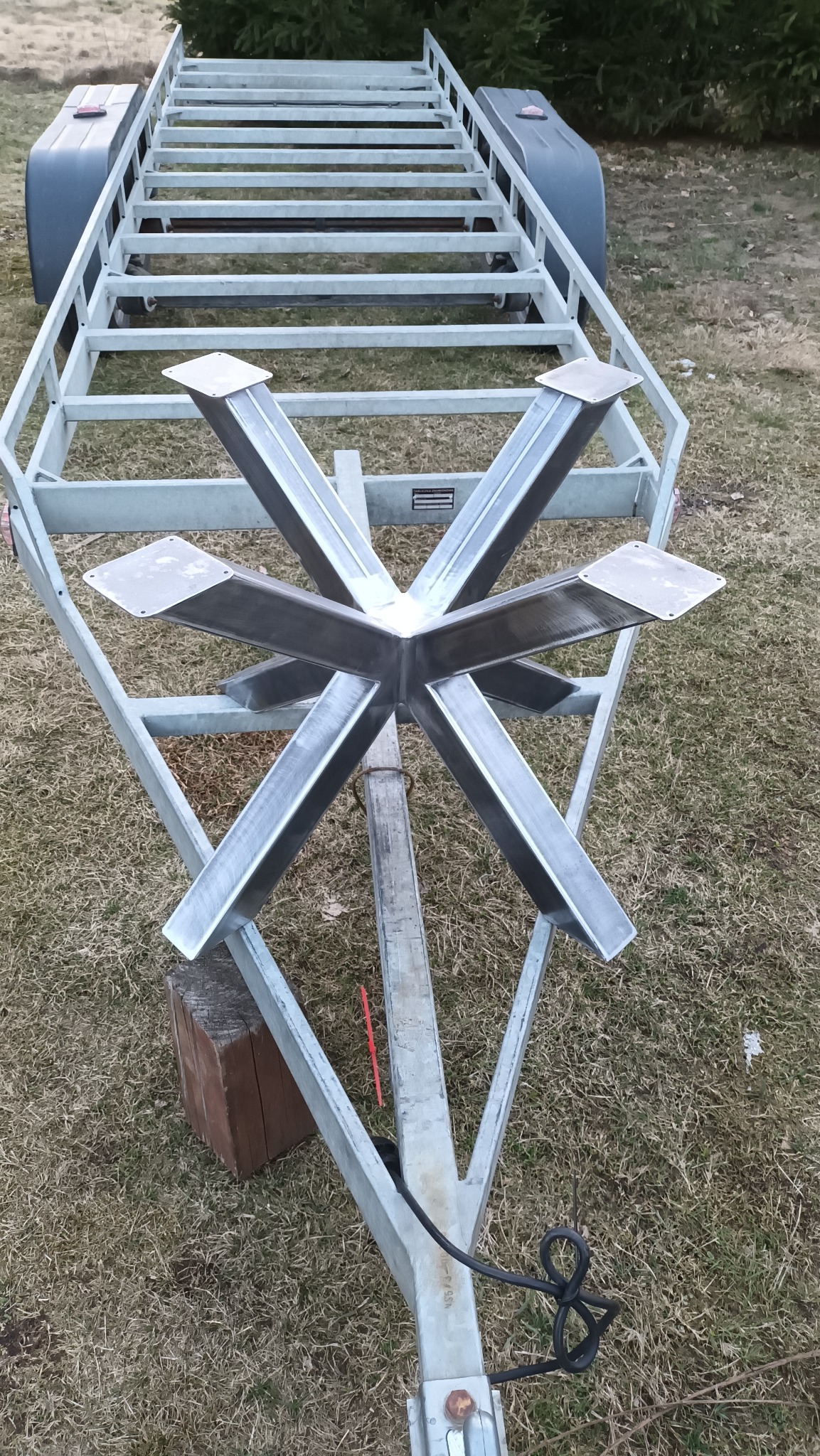 Metalowa konstrukcja przyczepy z elementami spawania, widoczna na tle trawy. Szczegółowy widok na łączenia i wsporniki. Nietypowy układ konstrukcyjny.