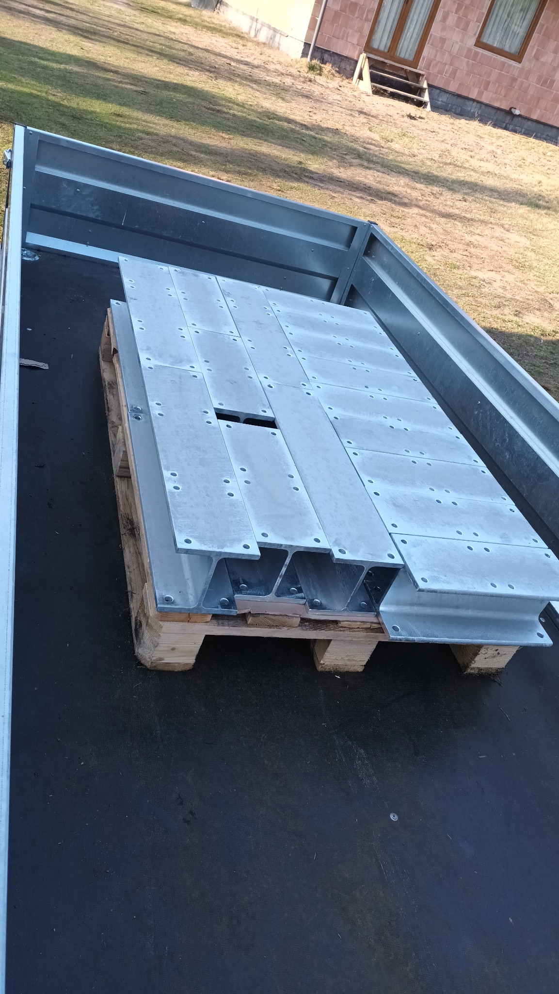 Stalowe elementy konstrukcyjne na palecie w przyczepie, przygotowane do montażu tarasu drewnianego na zewnątrz budynku.