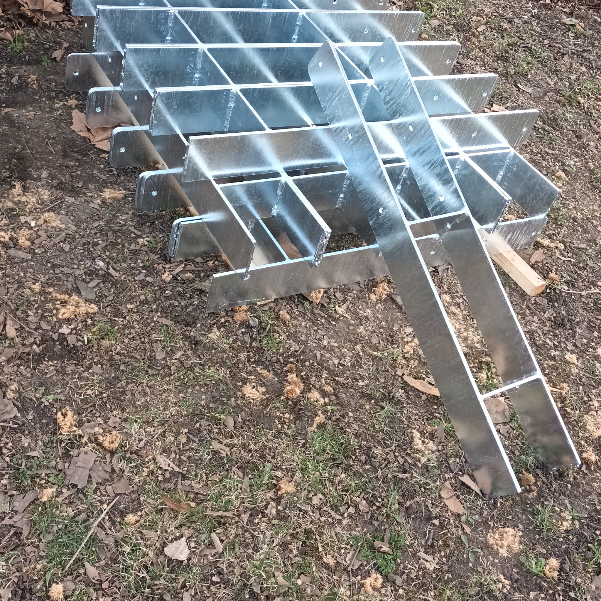 Stalowe, ocynkowane elementy konstrukcyjne, prawdopodobnie do budowy tarasu drewnianego, ułożone na ziemi. Widoczne otwory montażowe.