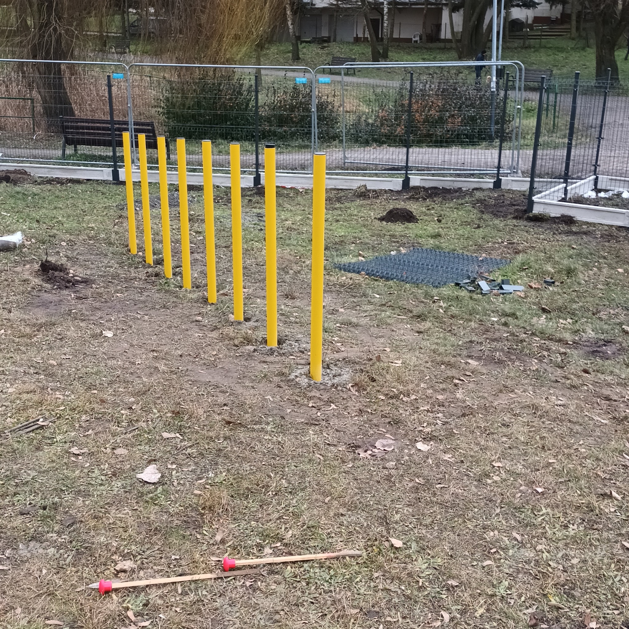 Żółte słupki w rzędzie na placu zabaw, elementy konstrukcyjne, teren przygotowany pod montaż, widoczne narzędzia i fragmenty nawierzchni.