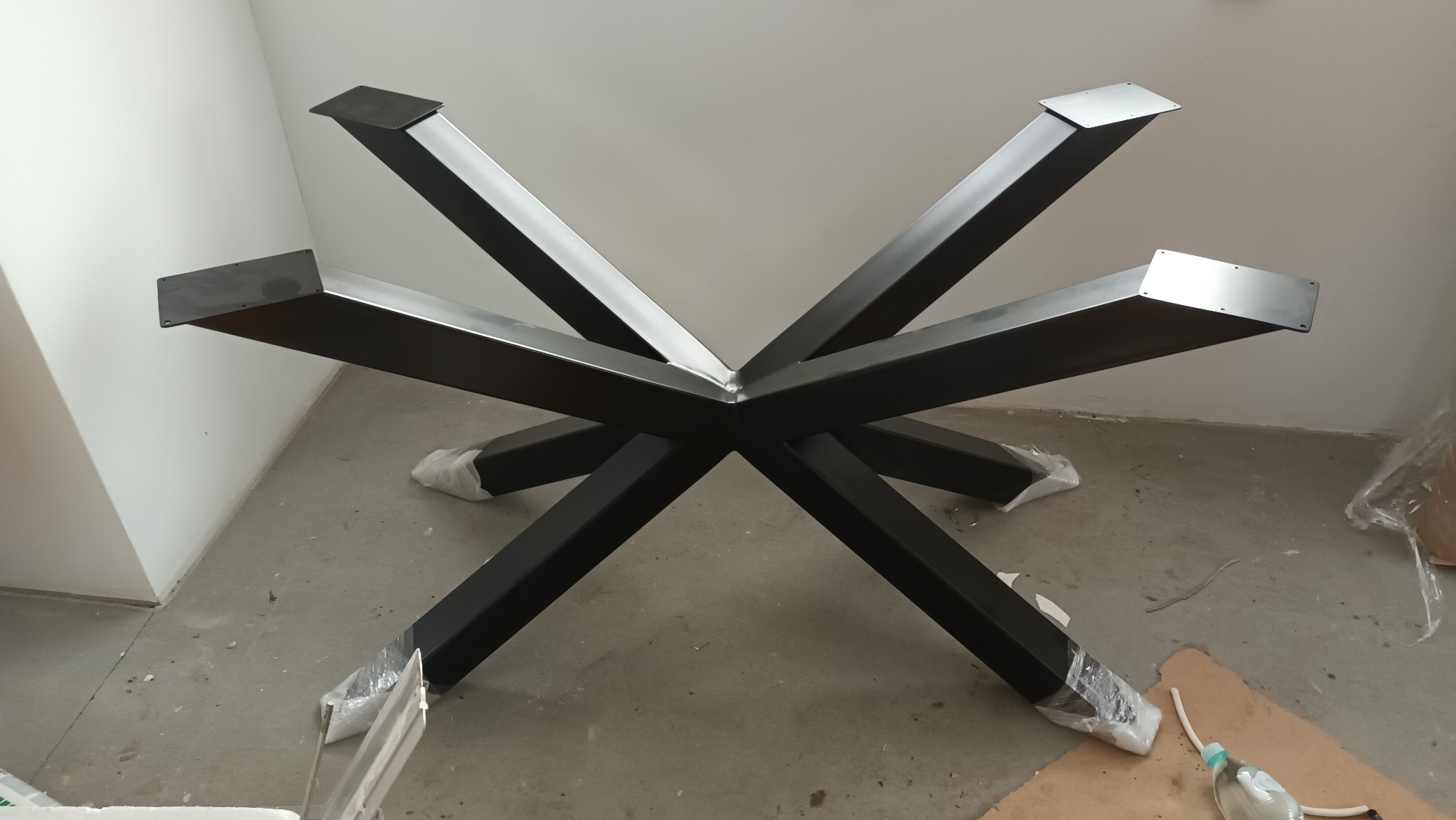 Metalowa podstawa stołu z czarnymi, skrzyżowanymi nogami, przygotowana do montażu blatu. Nogi zabezpieczone folią. Widok z góry, surowe wnętrze.