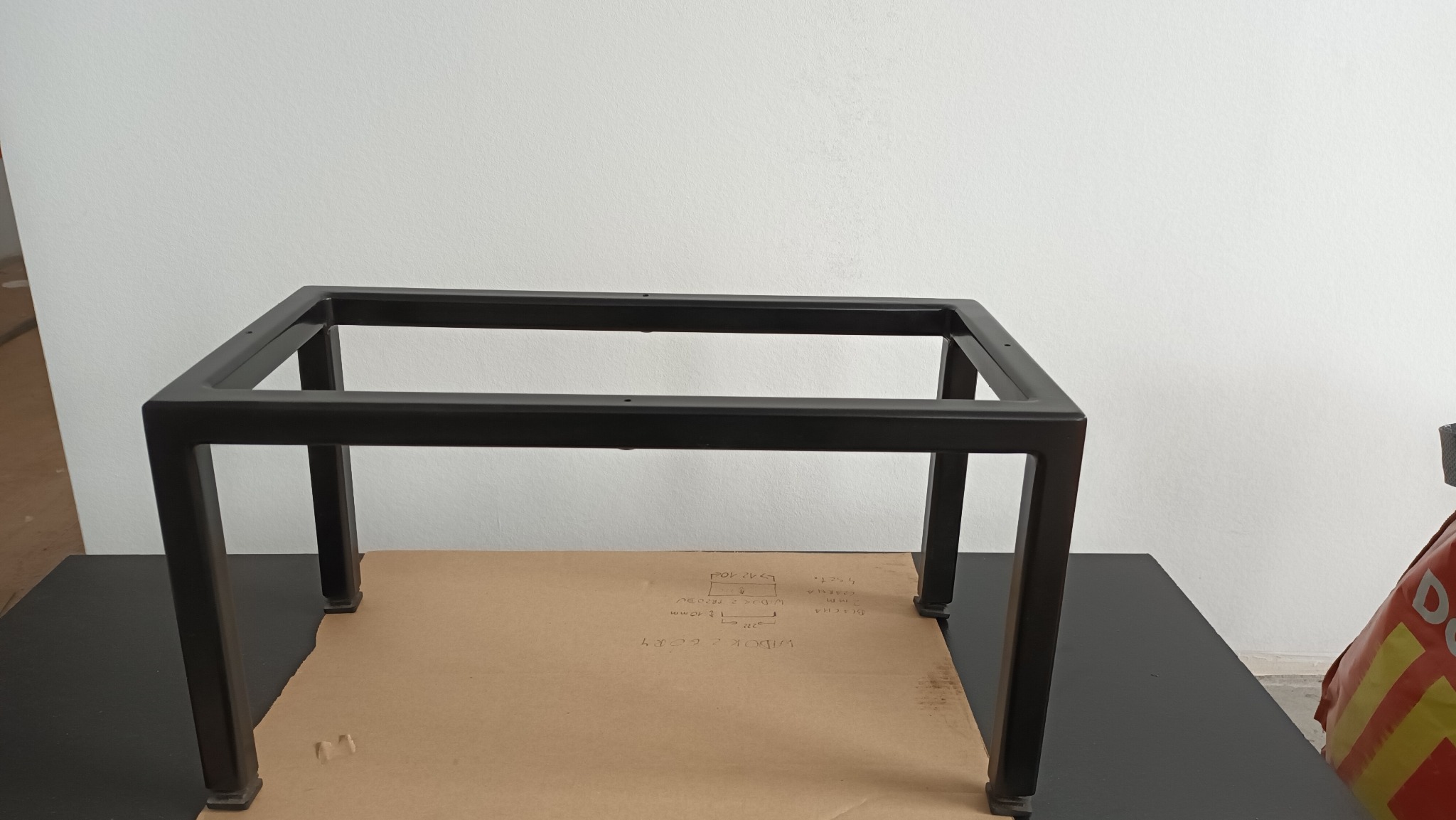 Czarna, metalowa konstrukcja stolika na kartonie z odręcznymi notatkami, na tle białej ściany. Widoczne elementy montażowe i fragment podłogi. Minimalistyczny design.