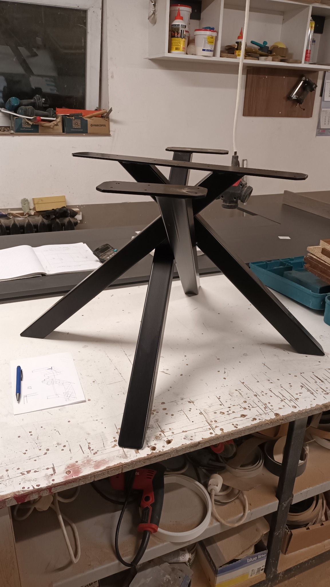 Metalowa, czarna podstawa stołu w warsztacie, widoczne narzędzia i rysunek techniczny. Cztery nogi rozchodzą się promieniście, na górze mocowania blatu.