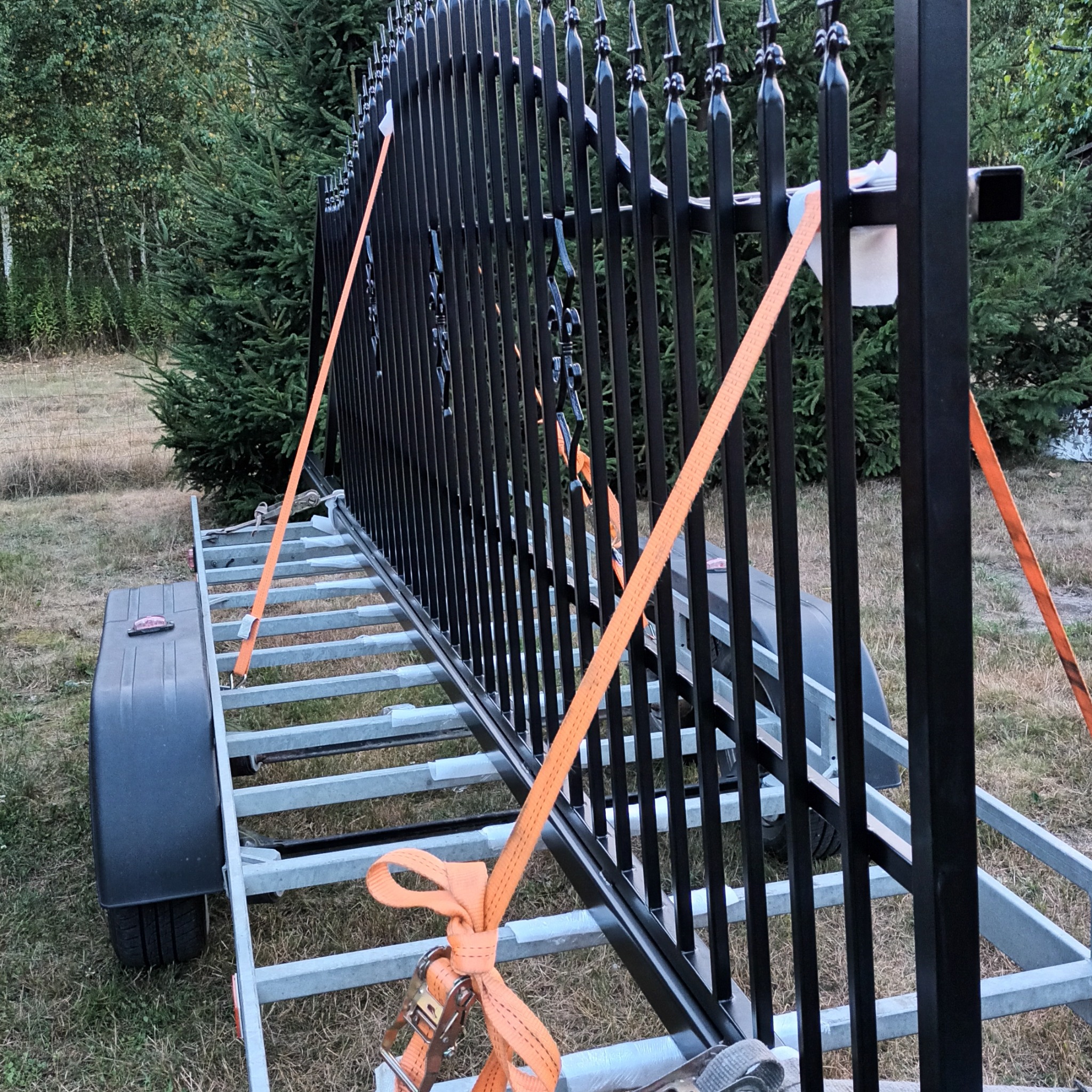 Czarny, metalowy płot z ozdobnymi grotami, zabezpieczony pomarańczowymi pasami transportowymi na przyczepce. W tle zieleń.