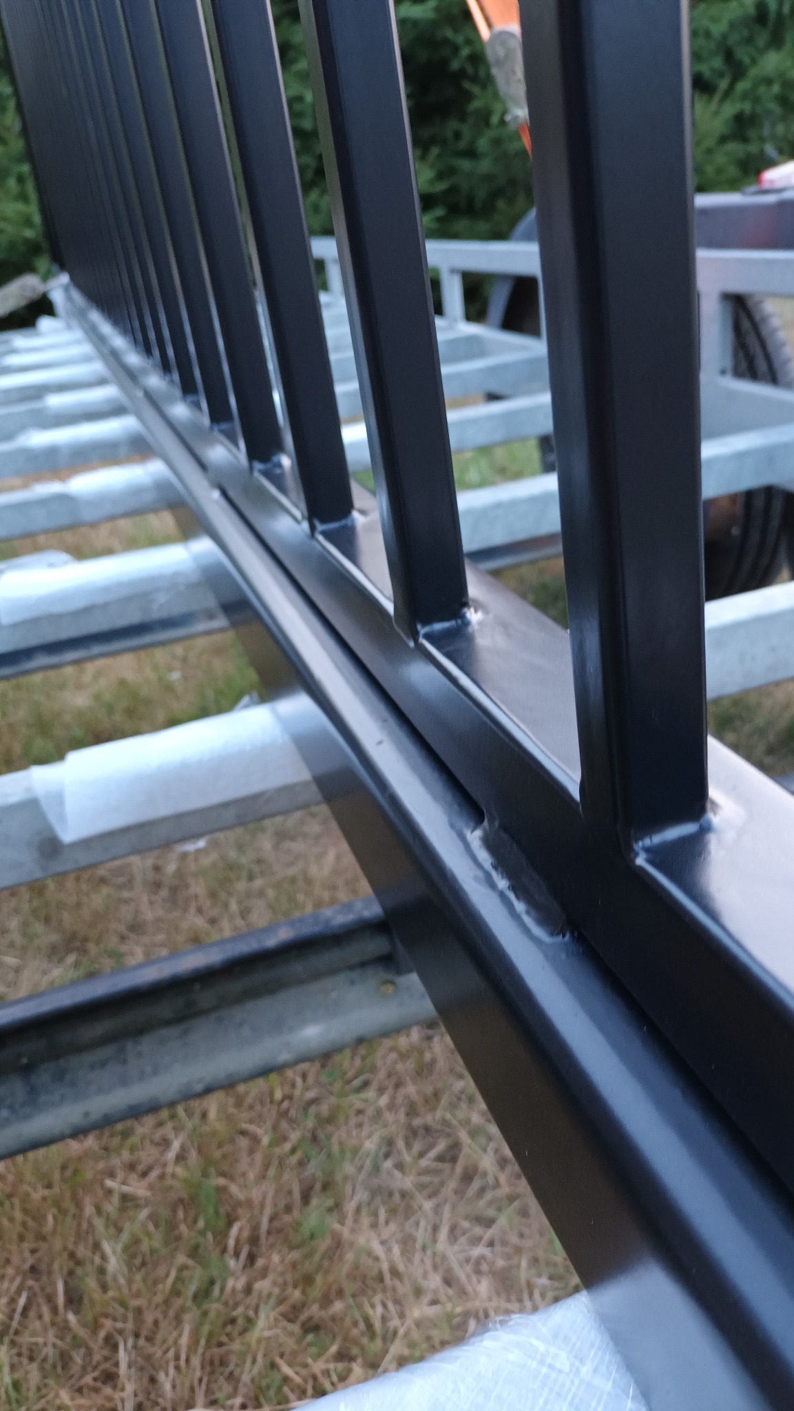 Czarny, metalowy płot z bliska, widoczne spawy i elementy konstrukcyjne. Tło z zielenią i fragmentem przyczepy. Detal ogrodzenia w trakcie montażu.