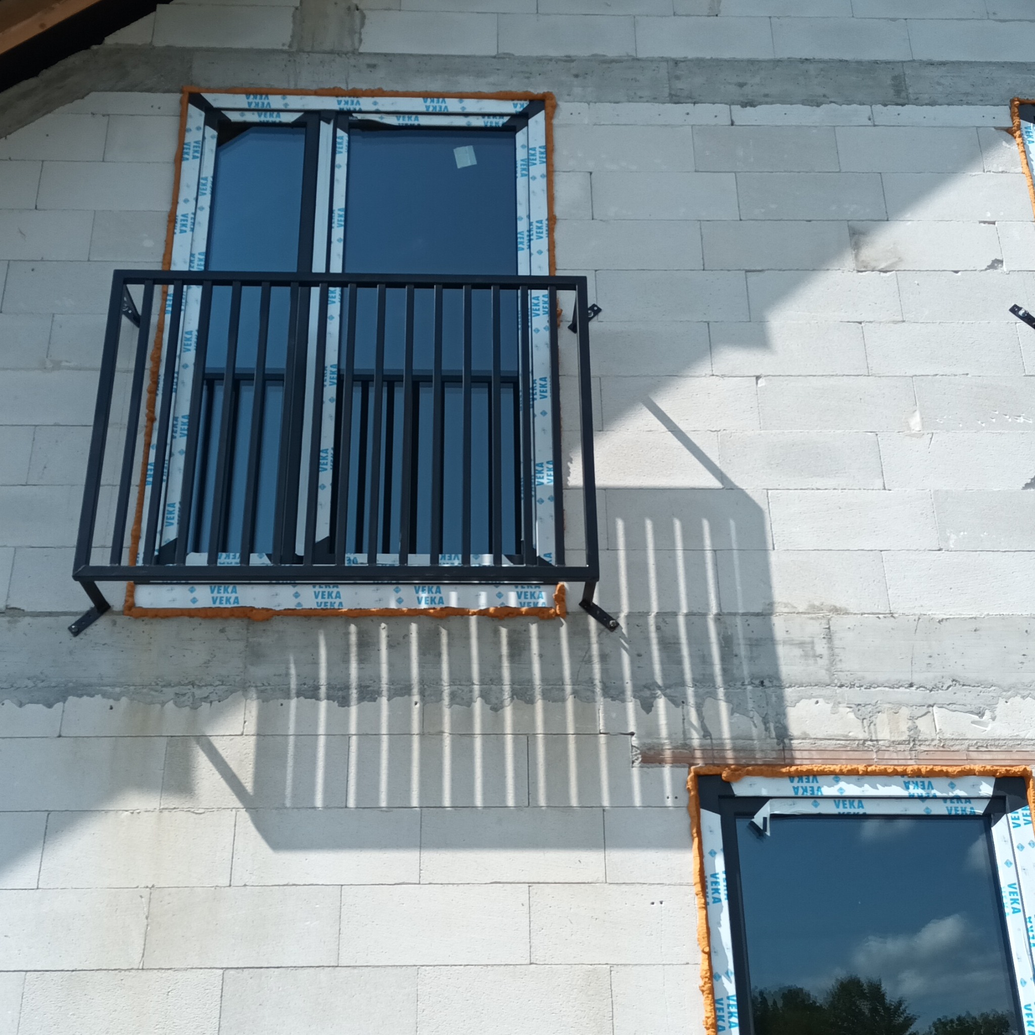 Nowy balkon z czarną balustradą przy oknie w surowym stanie budynku z białego bloczka. Widoczne piankowanie okna. Kątowe ujęcie z dołu, cień balustrady na ścianie.