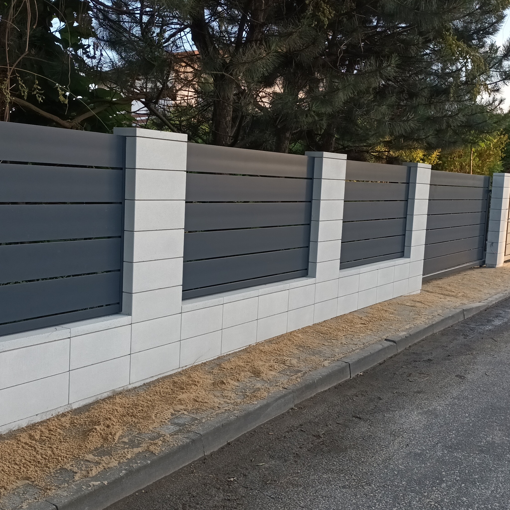 Nowoczesne ogrodzenie posesji: szare, poziome panele między białymi, kwadratowymi słupkami z bloczków. Widok na ulicę i drzewa w tle.