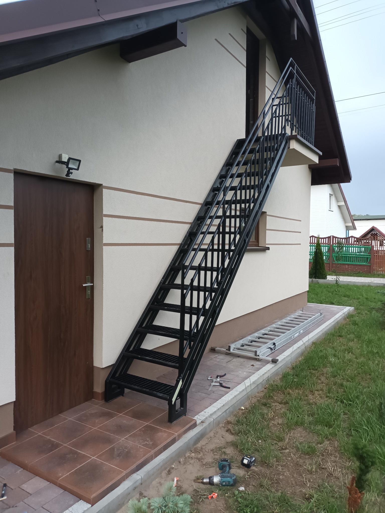 Zewnętrzne, metalowe schody prowadzące na balkon, przymocowane do ściany budynku z brązowymi drzwiami. Na ziemi narzędzia i drabina.