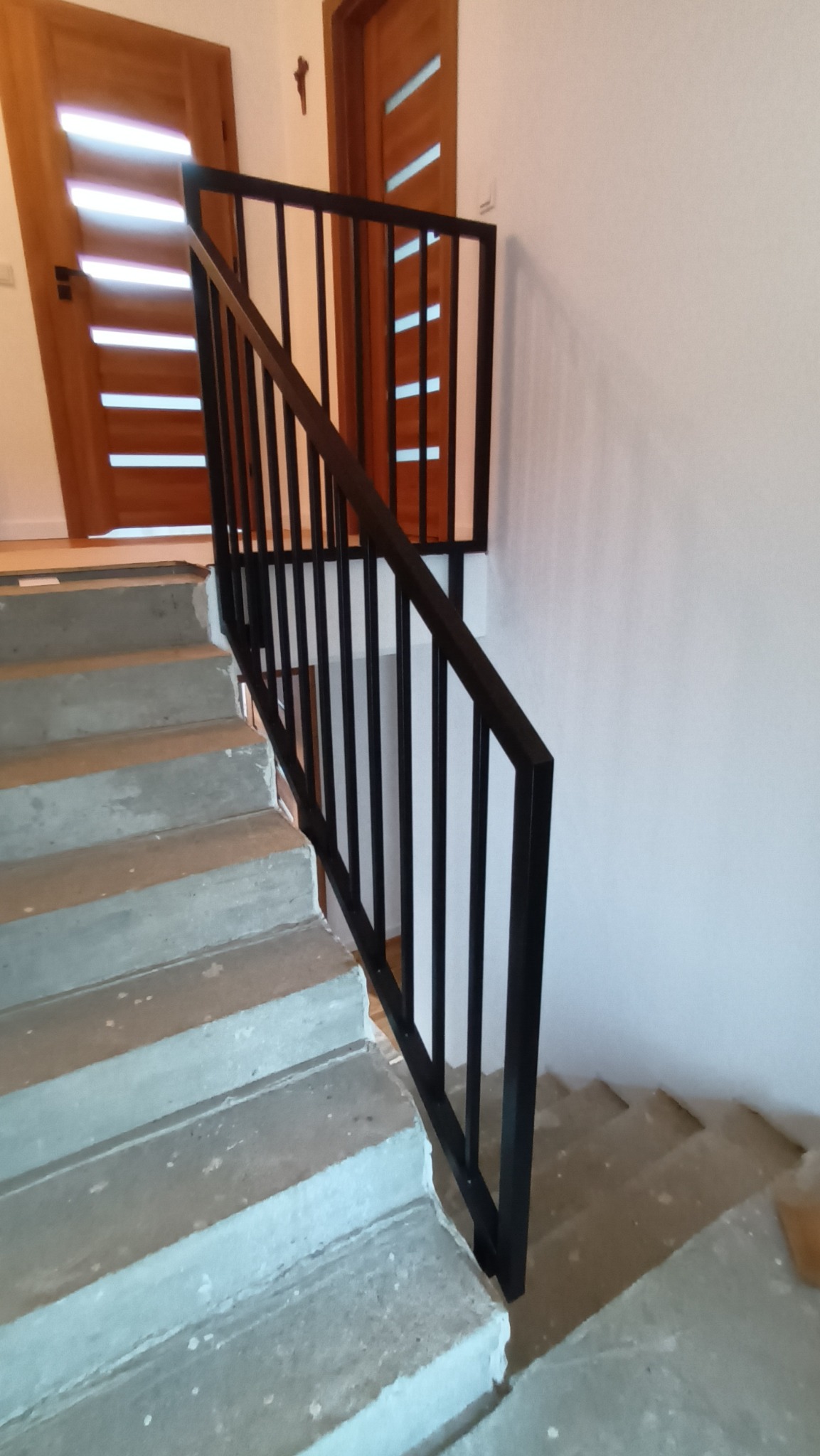 Nowoczesna, czarna balustrada schodowa z pionowymi szczeblami, zamontowana na surowych, betonowych schodach. W tle widoczne drewniane drzwi z przeszkleniami.