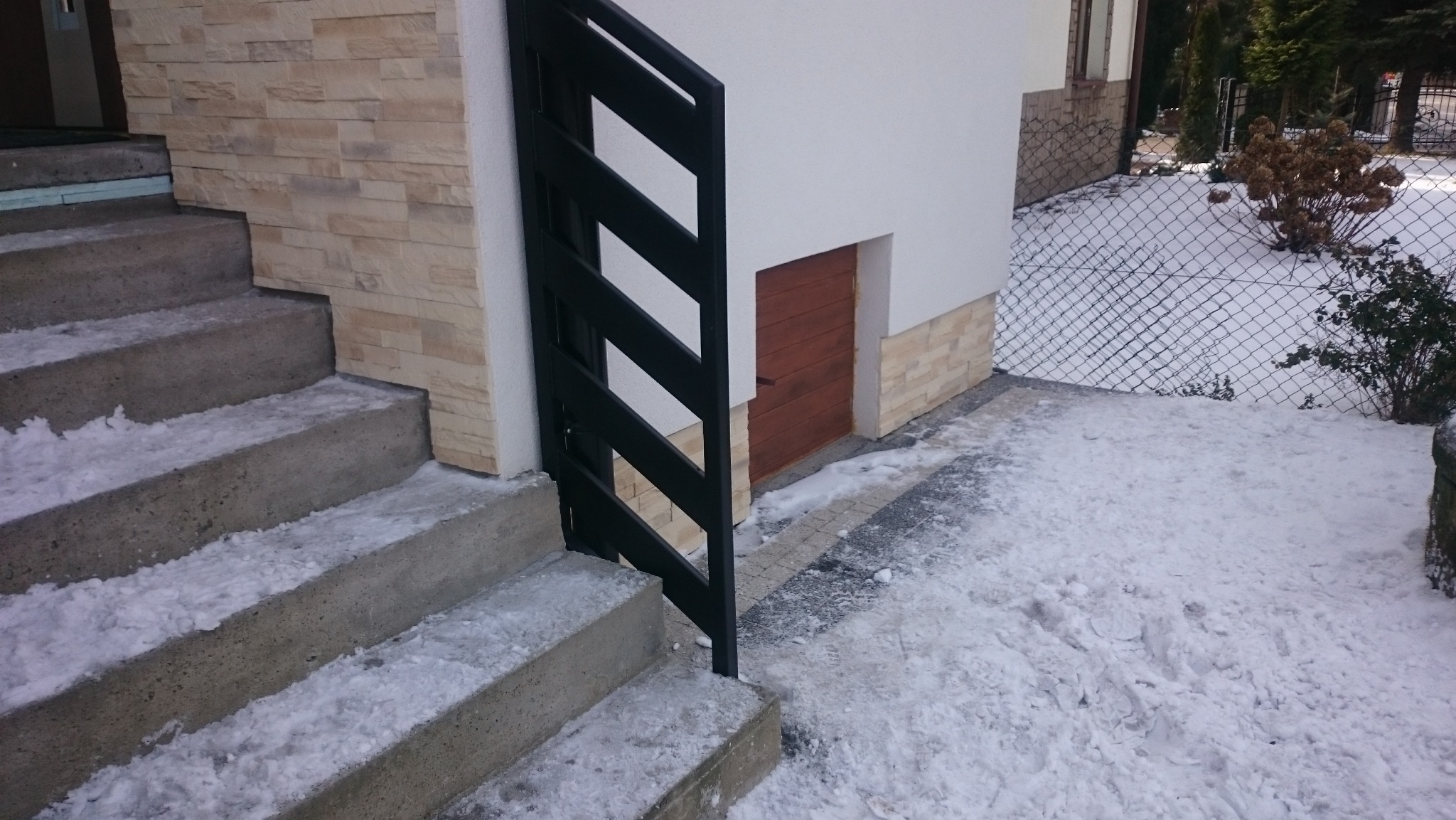 Betonowe schody z czarną balustradą prowadzą do budynku z elewacją z kamienia. Zima, śnieg na schodach i w ogrodzie. Małe drewniane drzwi w ścianie.