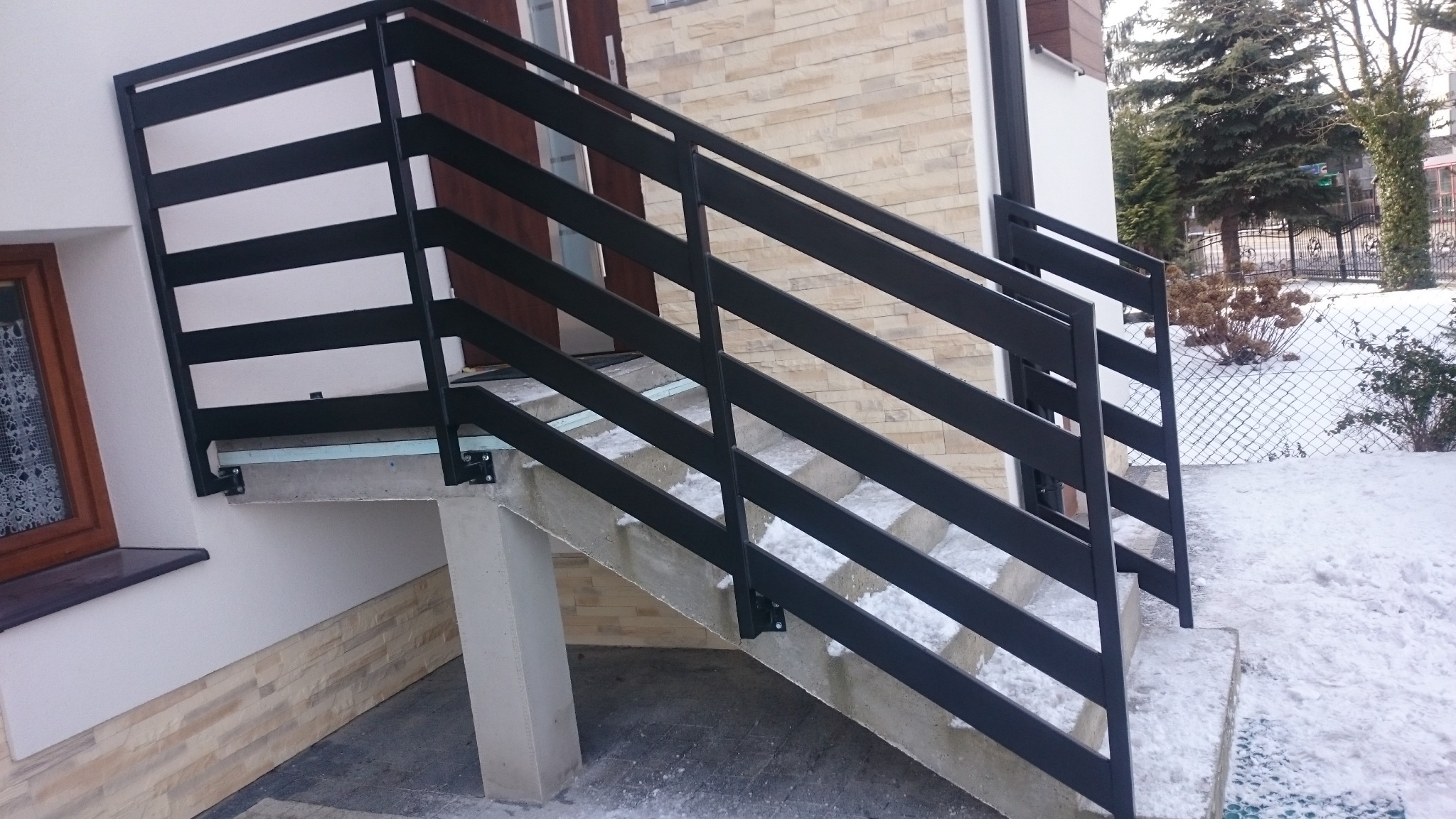 Zewnętrzne schody betonowe z czarną, metalową balustradą, prowadzące do drzwi wejściowych. W tle ośnieżony ogród i fasada budynku z dekoracyjną okładziną.
