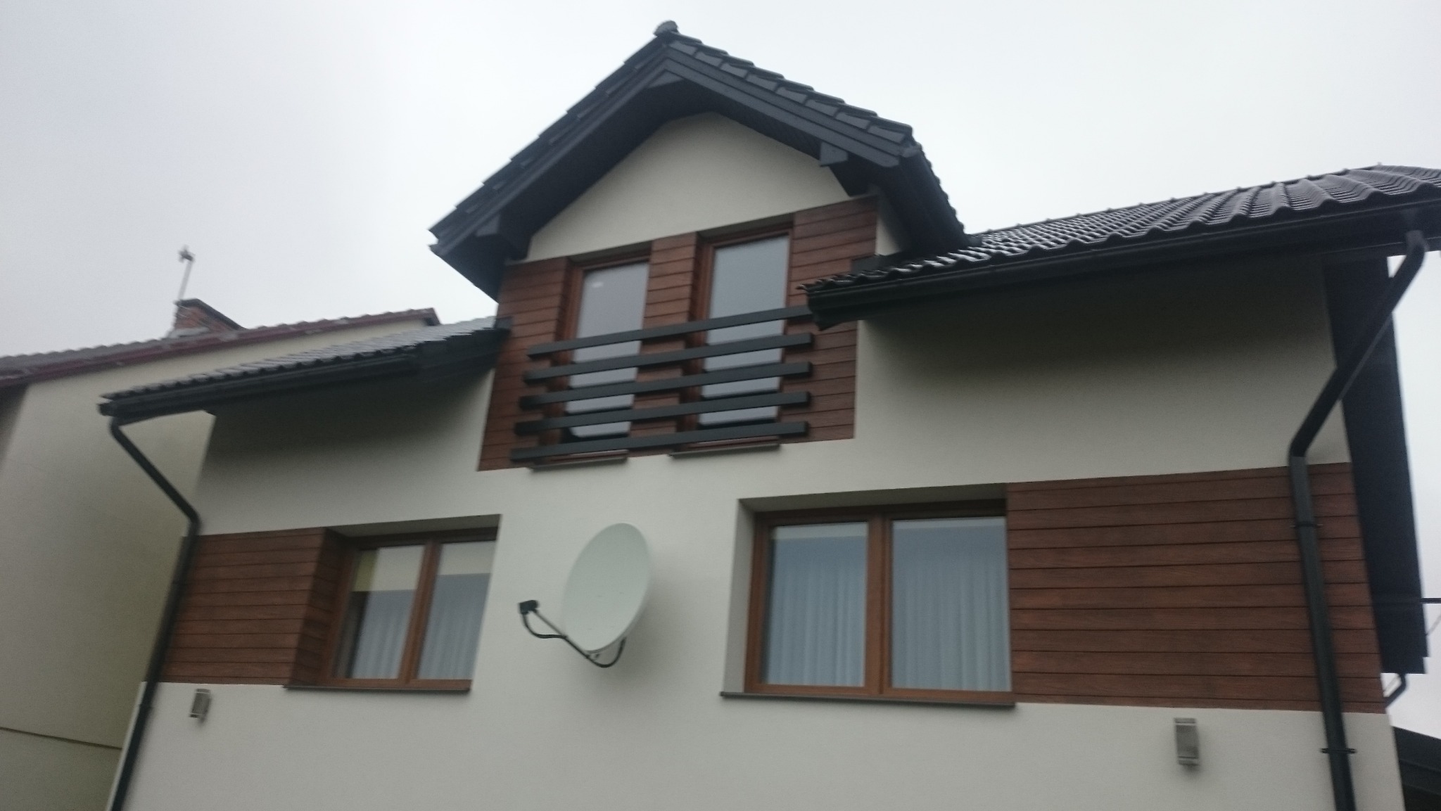 Elewacja domu z drewnianymi panelami i balkonem z czarną balustradą. Widoczna antena satelitarna. Ujęcie z dołu, pochmurne niebo w tle.
