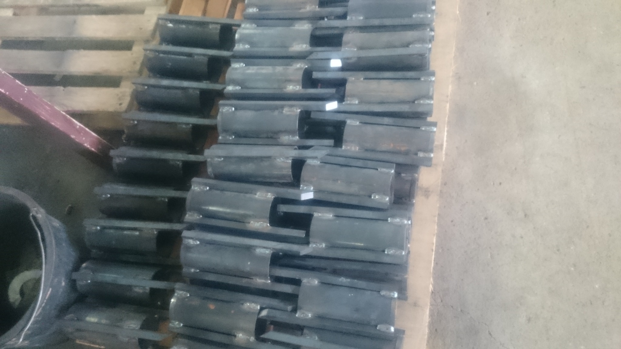 Rzędy spawanych, metalowych elementów konstrukcyjnych ułożonych na palecie, widoczne spoiny i niedoskonałości powierzchni. Elementy przygotowane do dalszej obróbki.