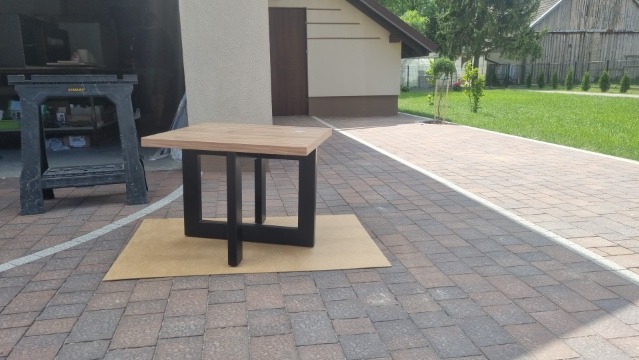 Kwadratowy, drewniany stolik z czarnymi nogami na kostce brukowej, na tle budynku gospodarczego i trawnika. Stolik leży na kartonie.