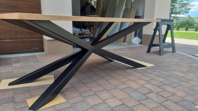 Stół z drewnianym blatem i metalową podstawą w kształcie gwiazdy, ustawiony na kostce brukowej. W tle widoczne narzędzia i materiały budowlane, co sugeruje montaż lub wykończenie.