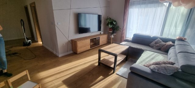 Jasne wnętrze salonu po remoncie: drewniana podłoga, szara sofa narożna, szafka RTV i TV na ścianie z dekoracyjnymi liniami. Widoczny odkurzacz w tle.