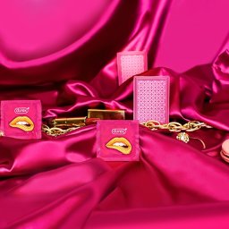 Katarzyna Fotorygo - Różowe opakowania prezerwatyw Durex z motywem ust, złoty łańcuch, karty do gry, szminka i makaroniki na błyszczącym, drapowanym, różowym materiale.