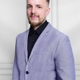 Katarzyna Fotorygo - Portret mężczyzny z brodą w czarnej koszuli i liliowym garniturze na tle białej ściany z panelami dekoracyjnymi.