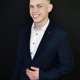 Katarzyna Fotorygo - Uśmiechnięty młody mężczyzna w granatowej marynarce i białej koszuli na czarnym tle, z ręką na klapie marynarki, prezentujący profesjonalny wizerunek.