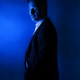 Katarzyna Fotorygo - Portret mężczyzny w tweedowej marynarce i kraciastej koszuli, oświetlonego intensywnym niebieskim światłem, spoglądającego przez ramię.
