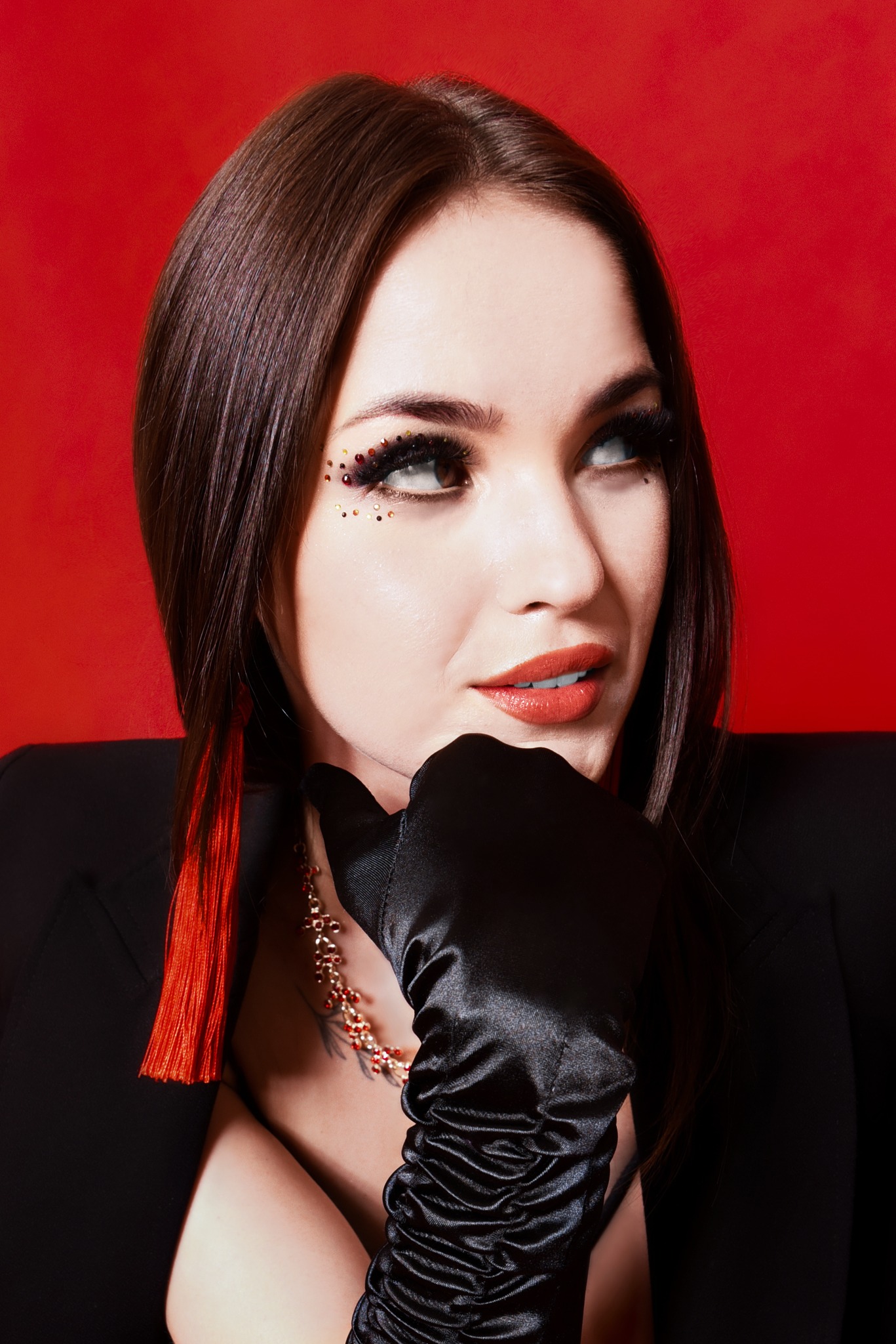 Portret kobiety w czarnym żakiecie i satynowych rękawiczkach, z ozdobami na twarzy i czerwonymi akcentami we włosach, pozującej na intensywnie czerwonym tle.