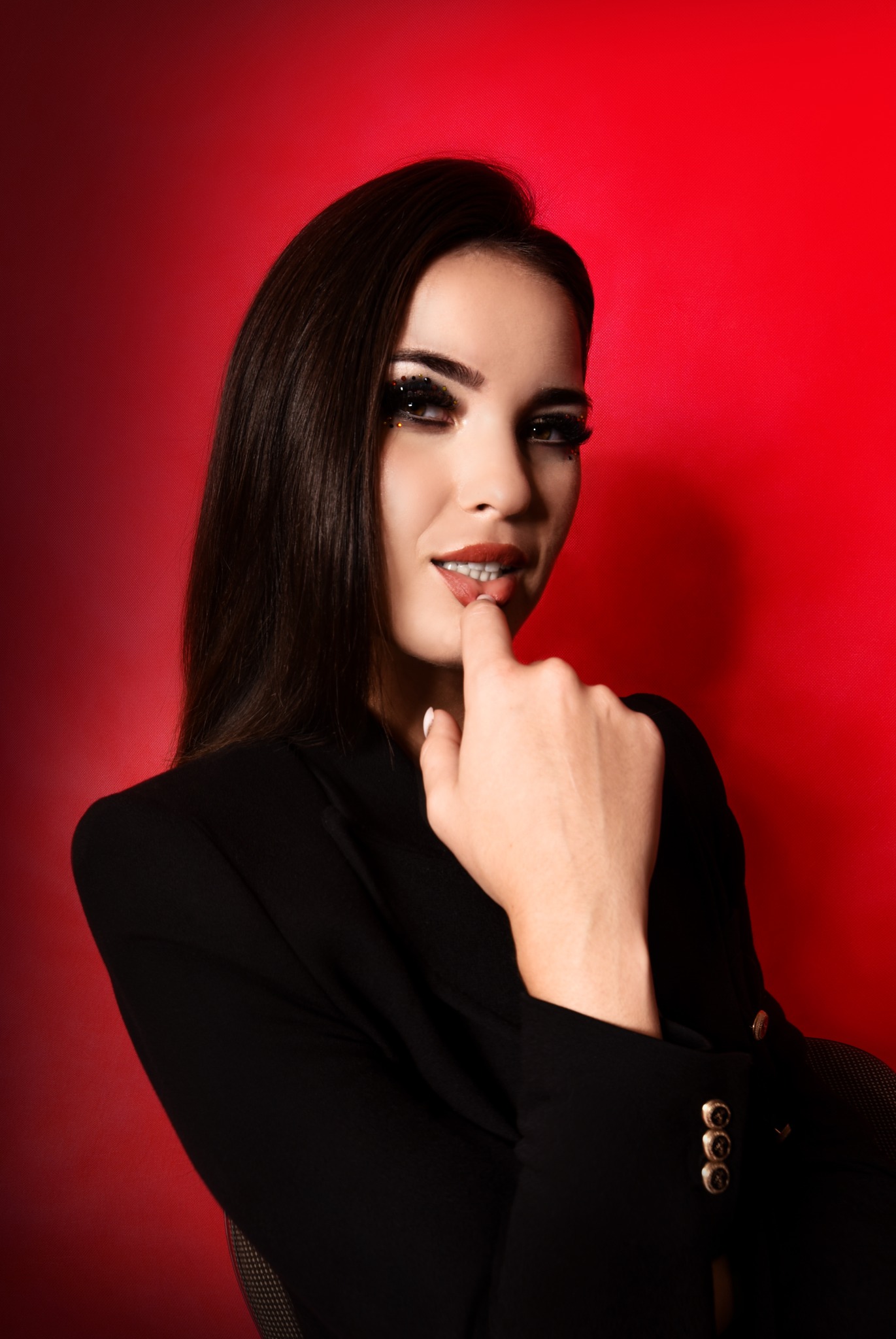 Portret kobiety w czarnej marynarce na czerwonym tle, z błyszczącym makijażem oczu i palcem przy ustach.
