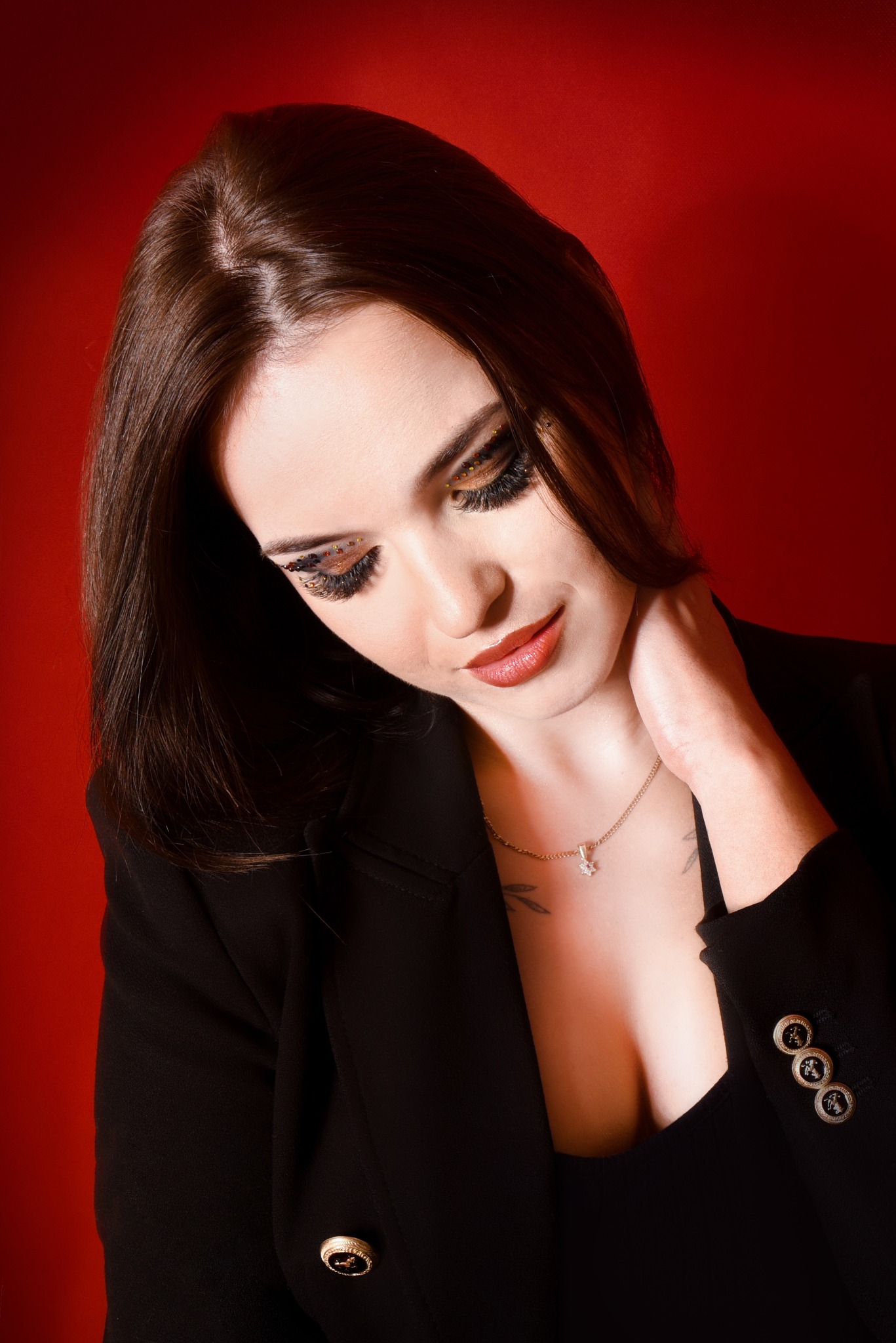 Portret kobiety w czarnej marynarce na czerwonym tle, z wyrazistym makijażem oczu z brokatem i pełnymi ustami, noszącej delikatny naszyjnik z wisiorkiem.