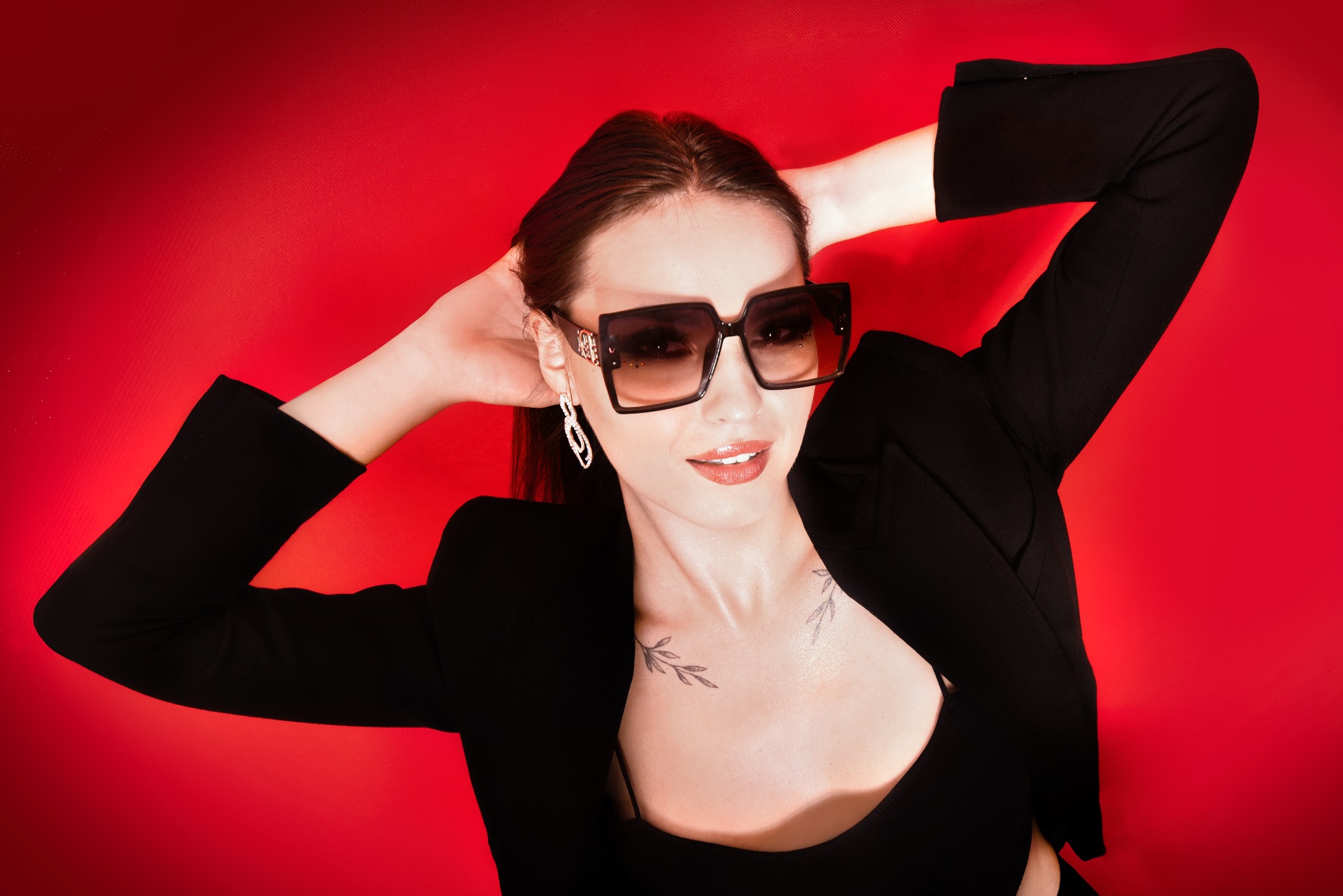 Portret kobiety w czarnym żakiecie i dużych okularach na czerwonym tle, z rękami uniesionymi za głową, eksponujący kolczyki i tatuaże na dekolcie.