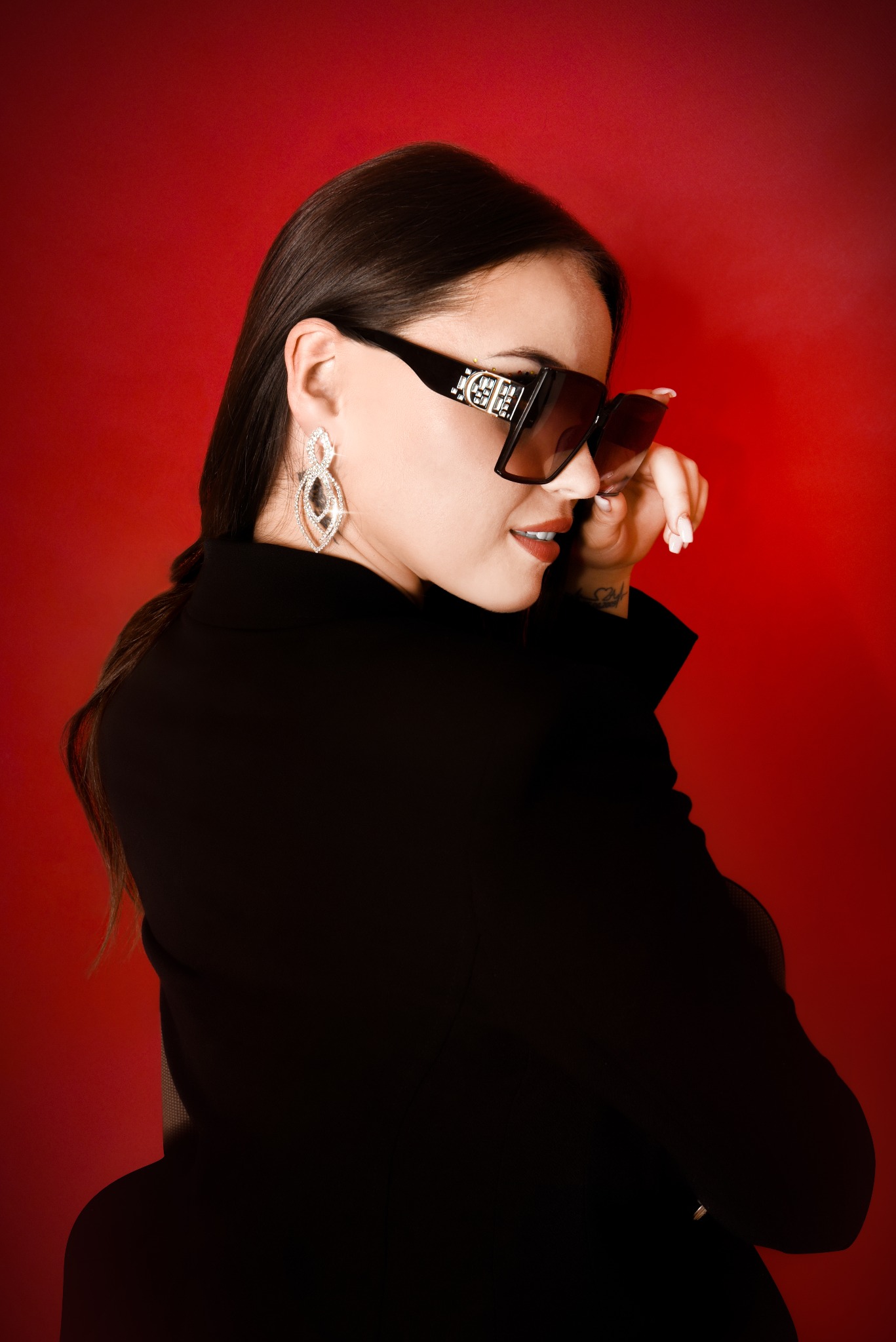 Portret kobiety w czarnej marynarce i dużych okularach przeciwsłonecznych z logo, na czerwonym tle, z widocznym tatuażem na nadgarstku i błyszczącymi kolczykami.