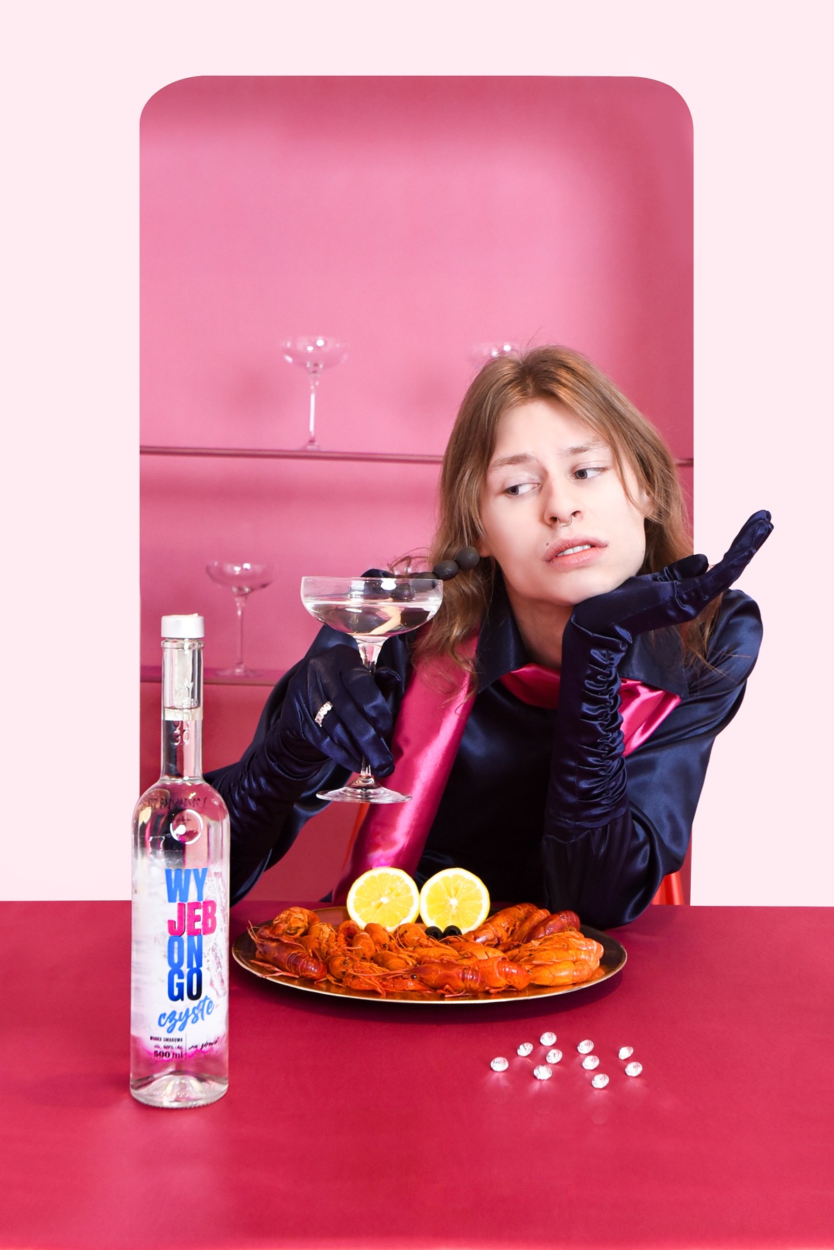 Osoba w satynowej bluzce i rękawiczkach pozuje z kieliszkiem koktajlowym, obok talerz z krewetkami i butelka wódki na różowym tle.