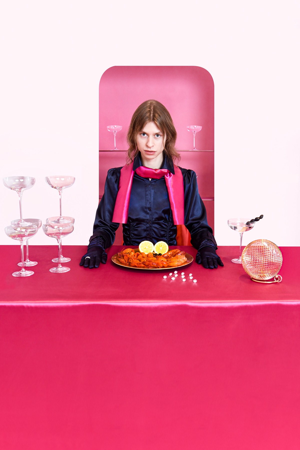 Kobieta w satynowej bluzce i rękawiczkach siedzi za stołem z krewetkami, cytryną i martini. W tle różowe tło z kieliszkami.