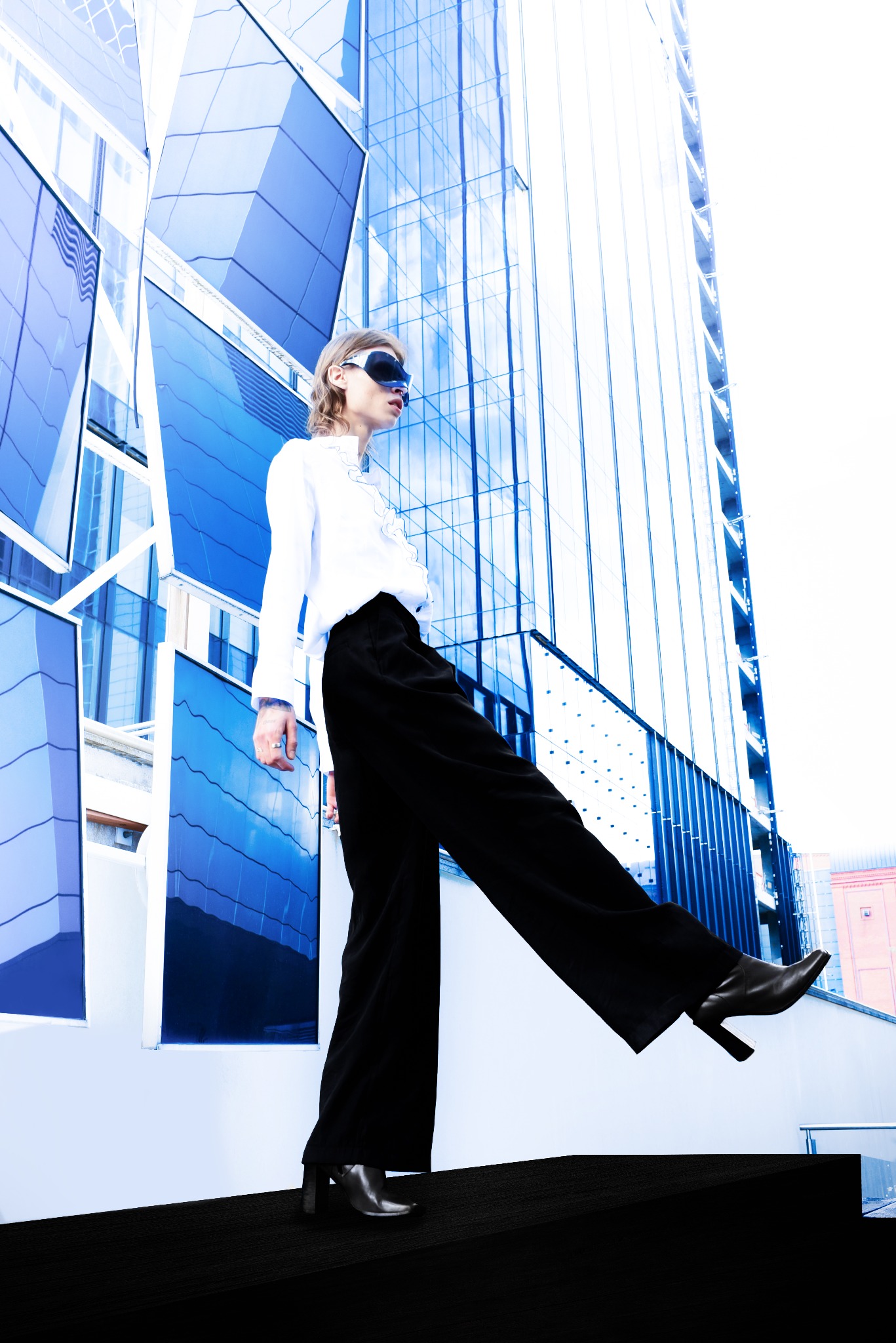 Osoba w białej koszuli i czarnych spodniach pozuje na tle nowoczesnego budynku z niebieskimi szklanymi panelami, nosząc futurystyczne okulary.