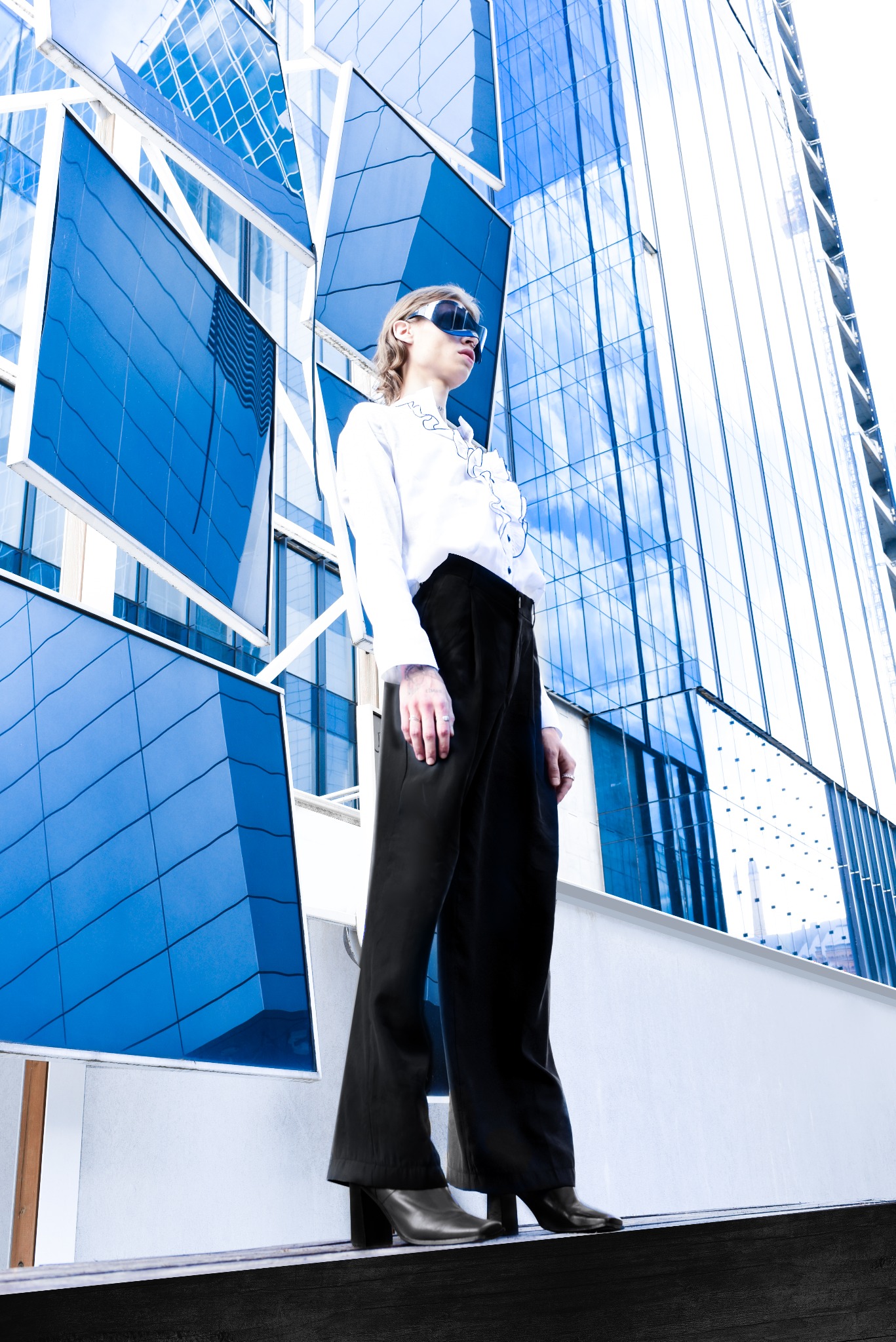 Osoba w okularach przeciwsłonecznych, białej koszuli i czarnych spodniach na tle nowoczesnego budynku z niebieskimi szklanymi panelami, ujęcie z dołu.