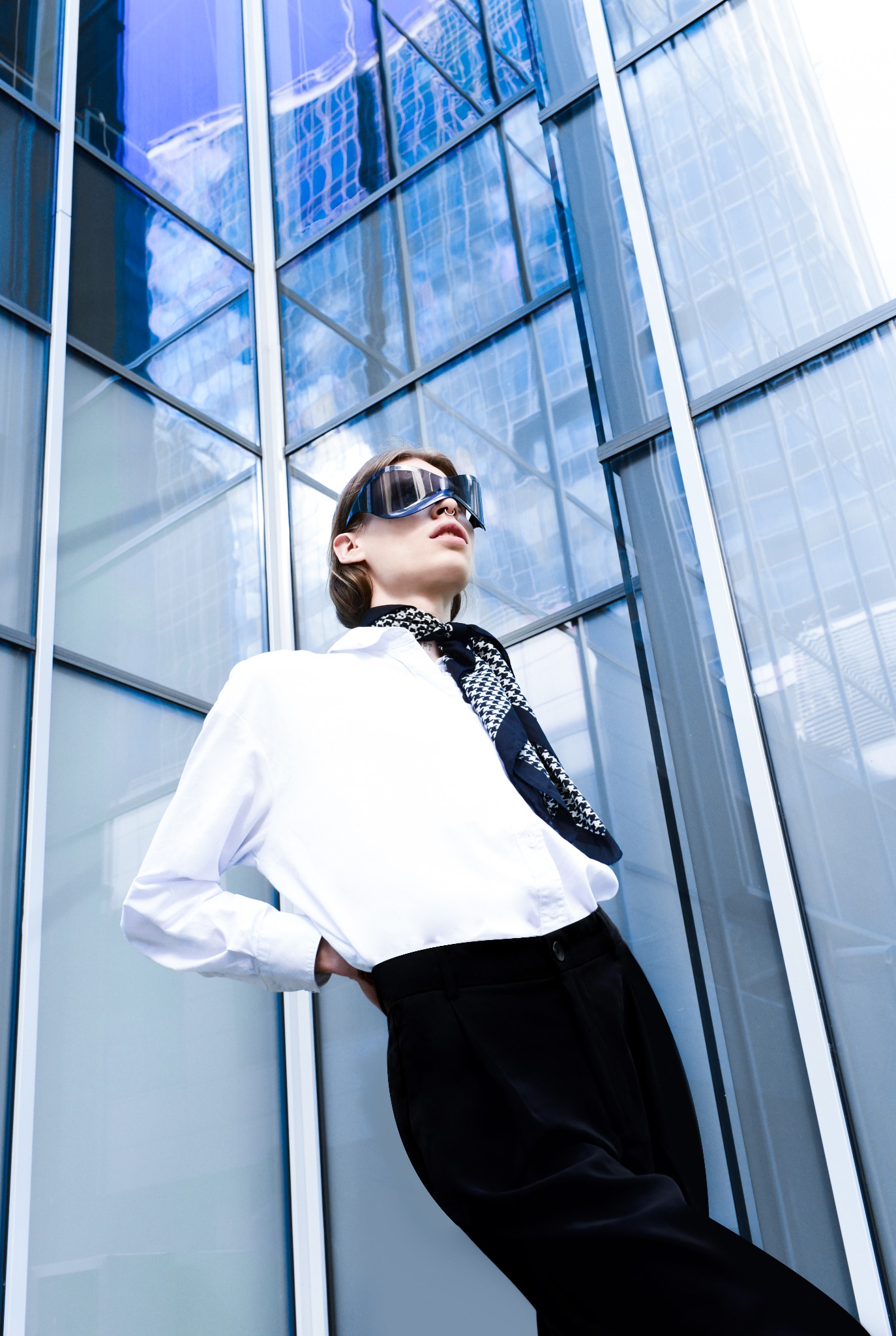 Młoda osoba w Poznaniu, ubrana w białą koszulę i czarne spodnie, pozuje na tle nowoczesnego, szklanego budynku, nosząc futurystyczne okulary i wzorzystą apaszkę.