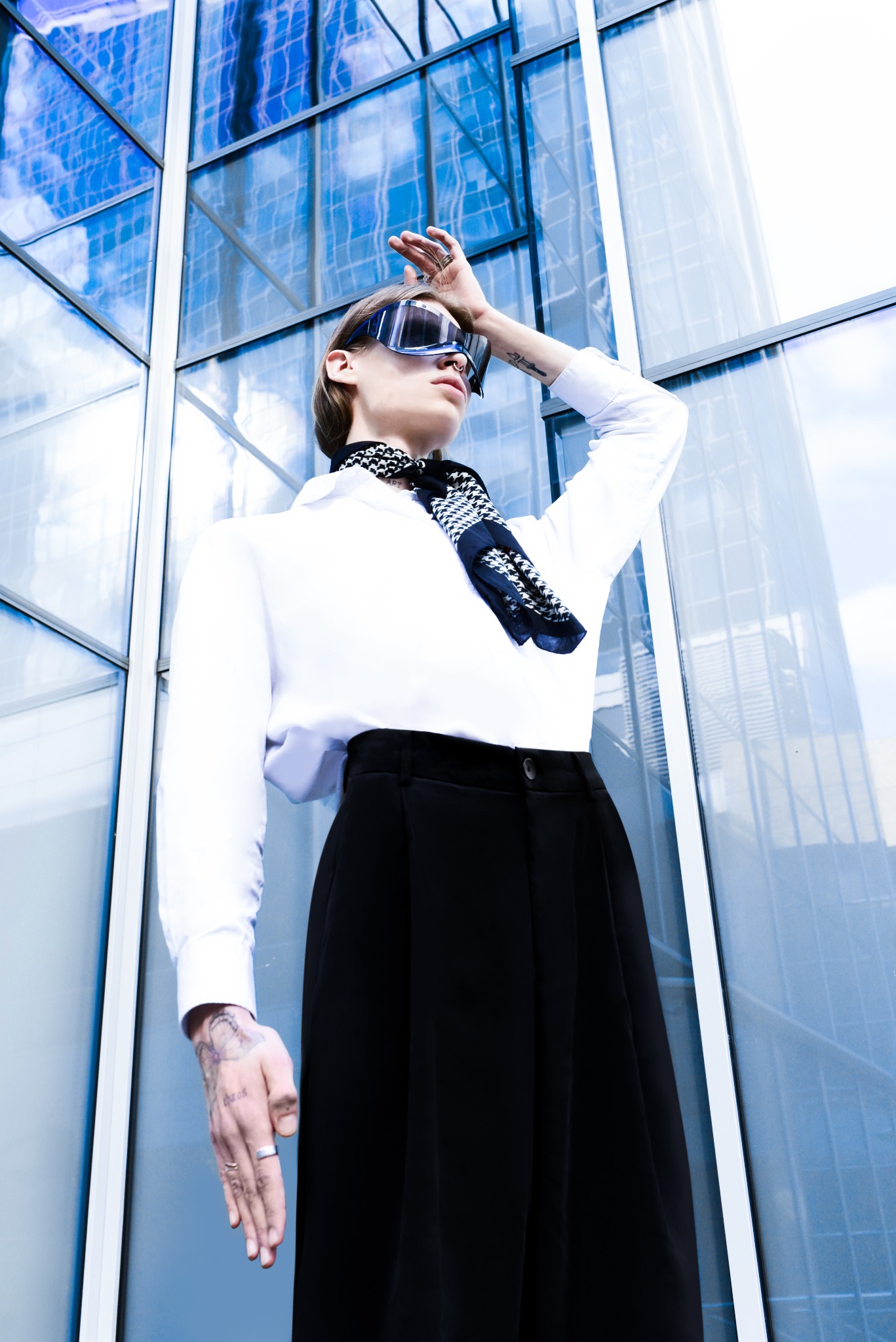 Osoba w białej koszuli i czarnej spódnicy, z wzorzystą apaszką na szyi i okularami przeciwsłonecznymi, pozuje na tle nowoczesnego budynku z szklaną fasadą, z widocznymi tatuażami na dłoniach...