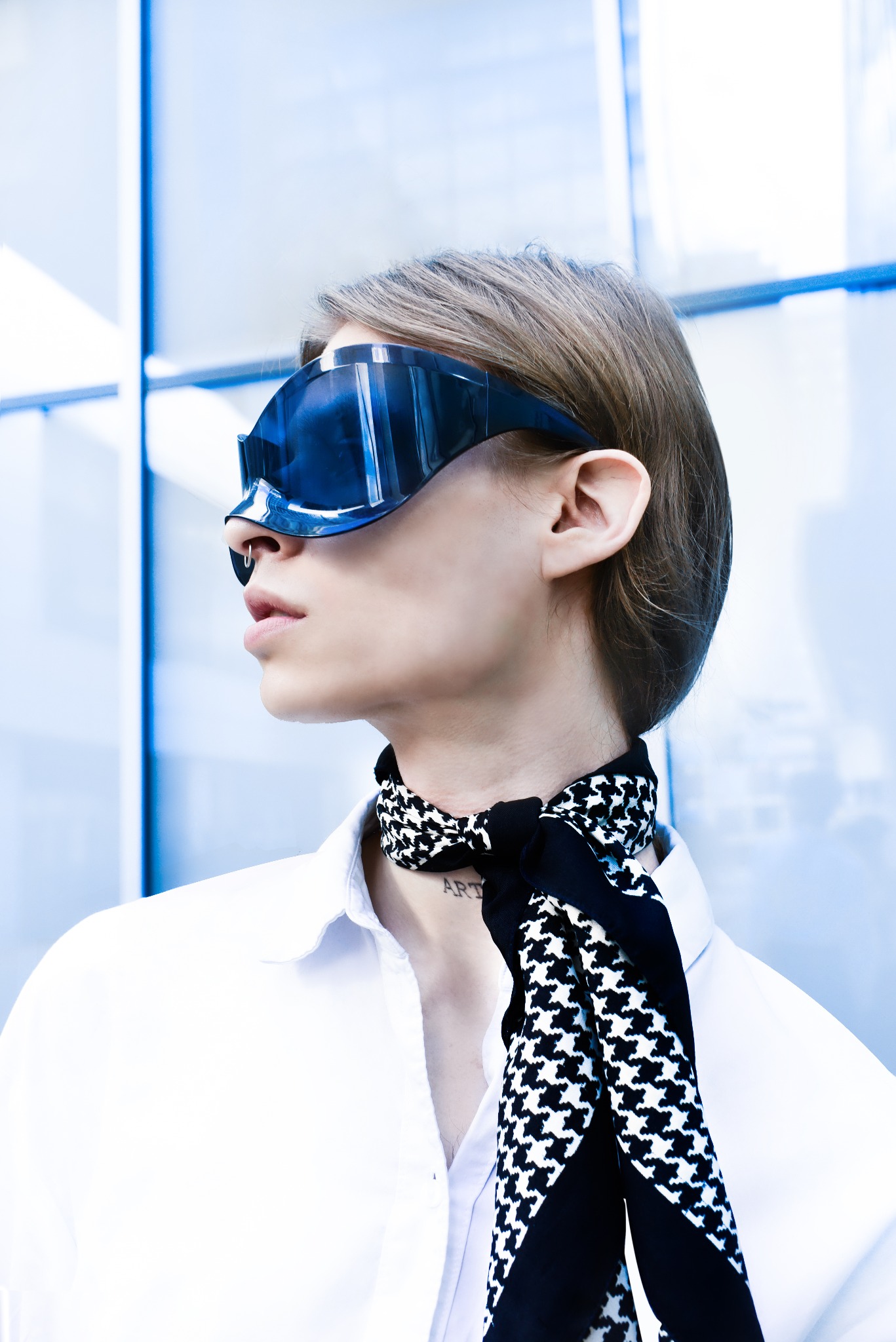 Portret osoby z futurystycznymi okularami przeciwsłonecznymi, białą koszulą i wzorzystą apaszką, stojącej na tle nowoczesnej, szklanej fasady.