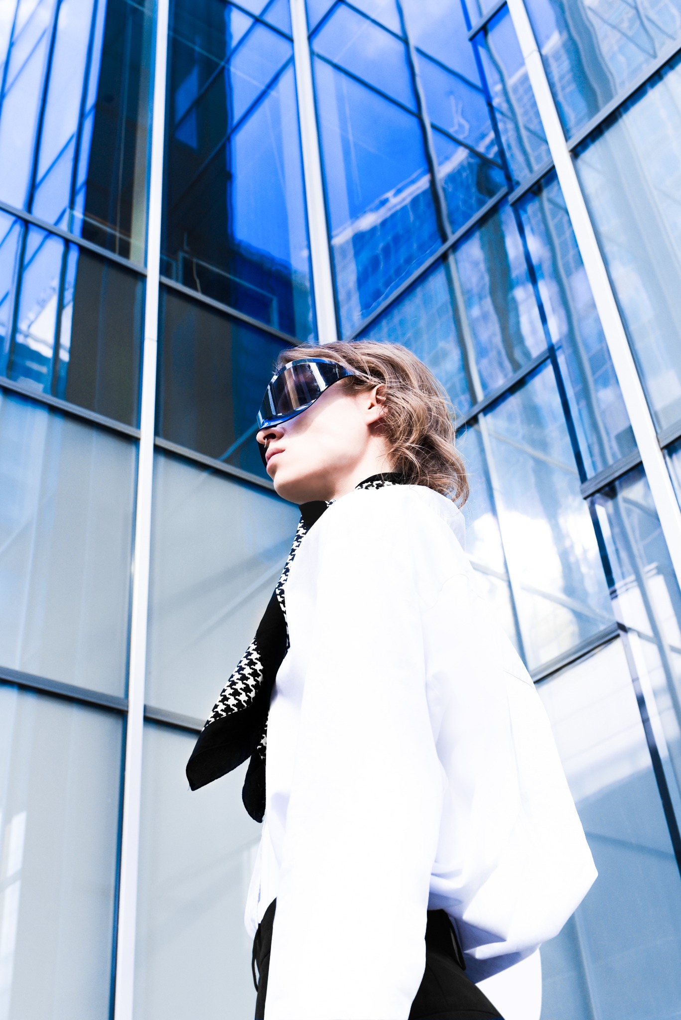 Osoba w okularach przeciwsłonecznych i białej koszuli na tle nowoczesnego budynku ze szklaną fasadą, ujęcie z dołu.