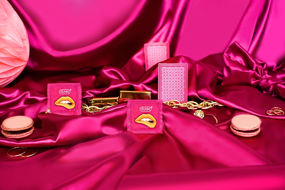 Różowe opakowania prezerwatyw Durex z motywem ust, złoty łańcuch, karty do gry, szminka i makaroniki na błyszczącym, drapowanym, różowym materiale.