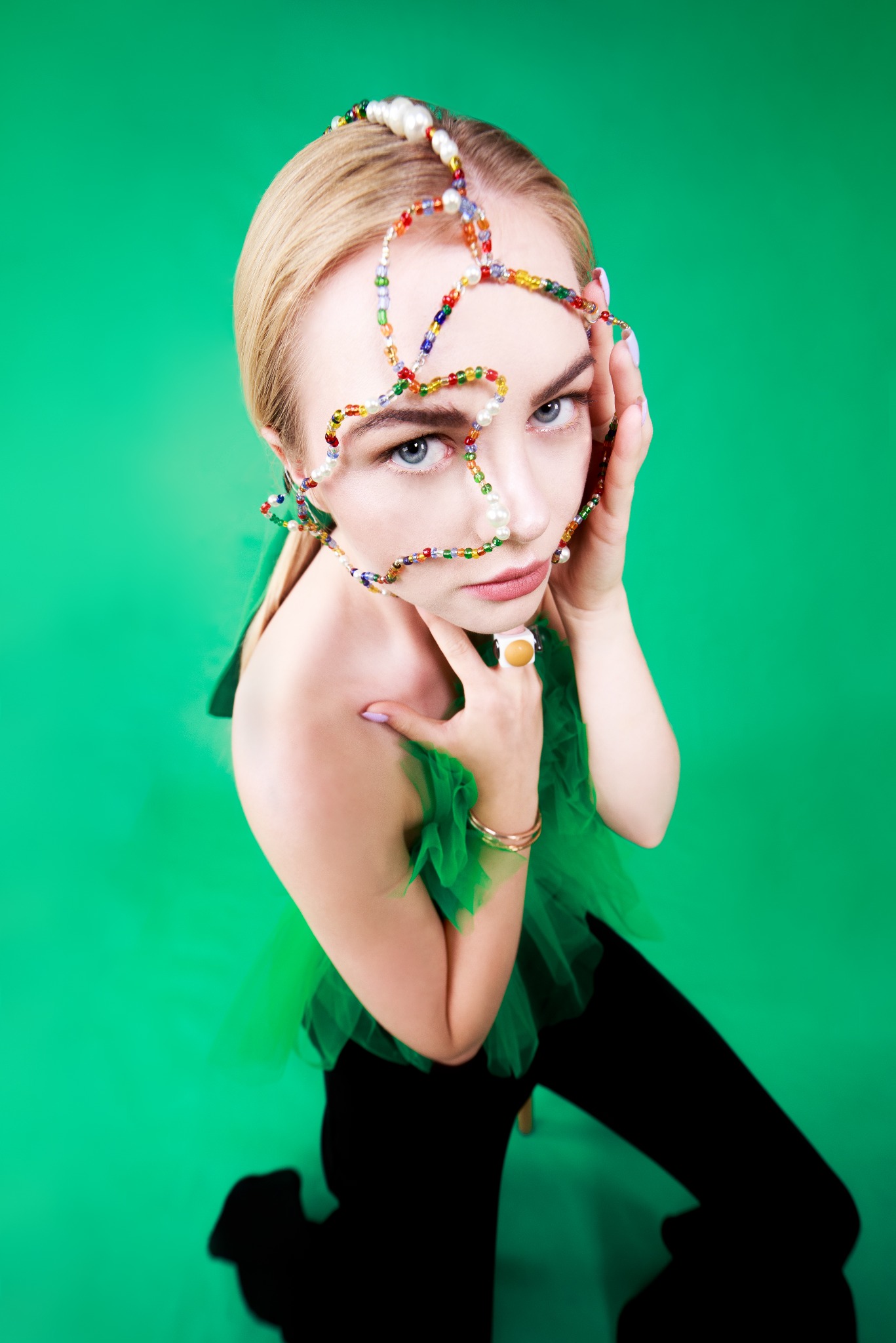 Portret kobiety z blond włosami i niebieskimi oczami na zielonym tle, z kolorowymi koralikami wokół twarzy i szyi, ubrana w tiulową bluzkę i czarne spodnie, z pierścionkiem na palcu.