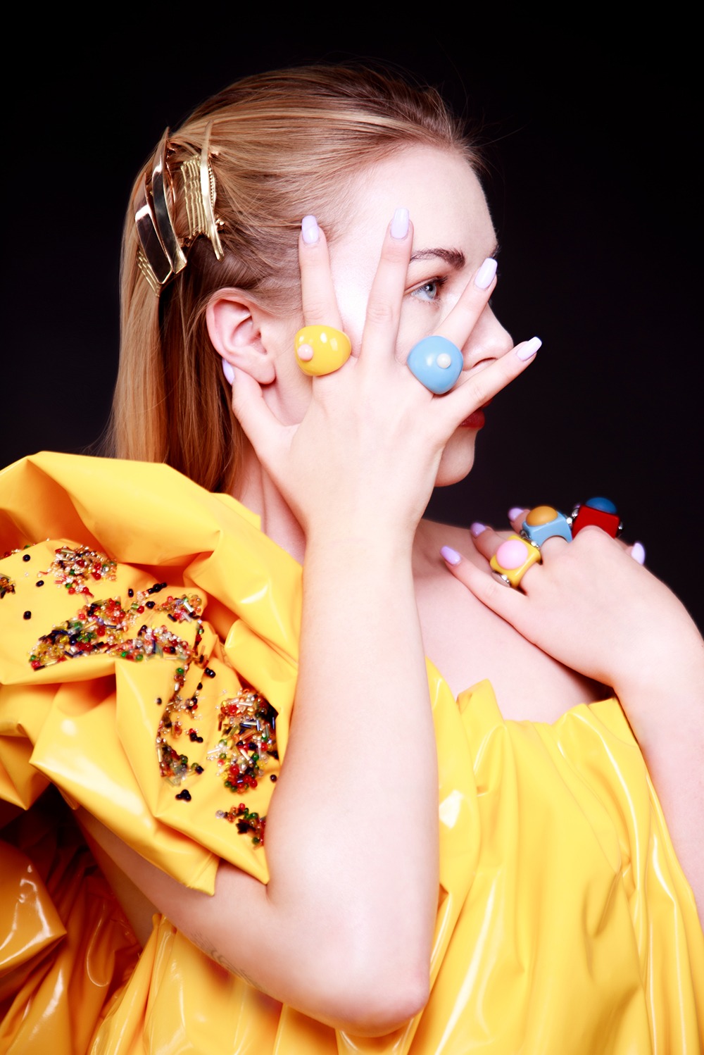 Portret kobiety w żółtej sukni z błyszczącymi aplikacjami, z geometryczną spinką we włosach i dużymi, kolorowymi pierścionkami na palcach, pozującej na czarnym tle.