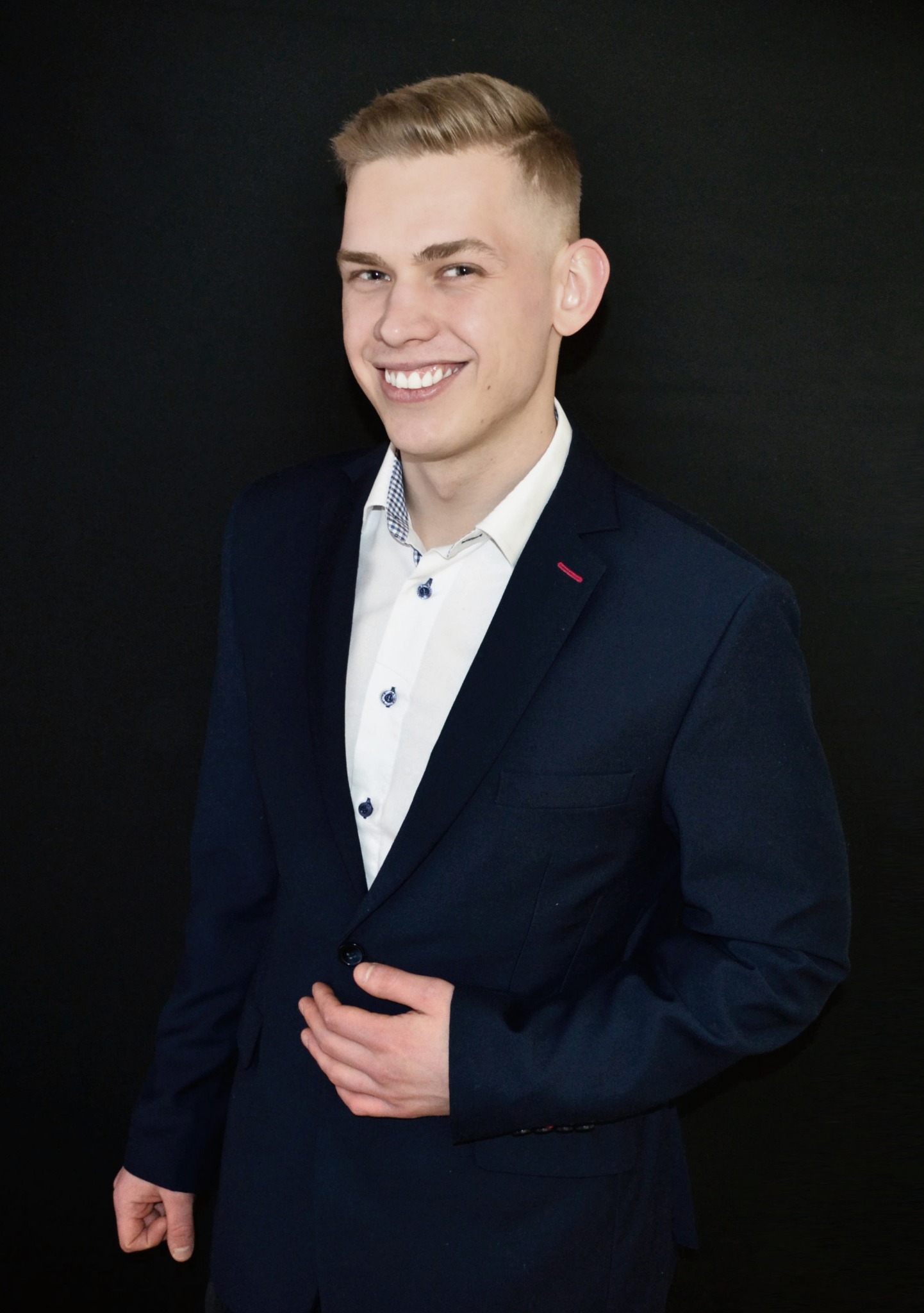 Uśmiechnięty młody mężczyzna w granatowej marynarce i białej koszuli na czarnym tle, z ręką na klapie marynarki, prezentujący profesjonalny wizerunek.