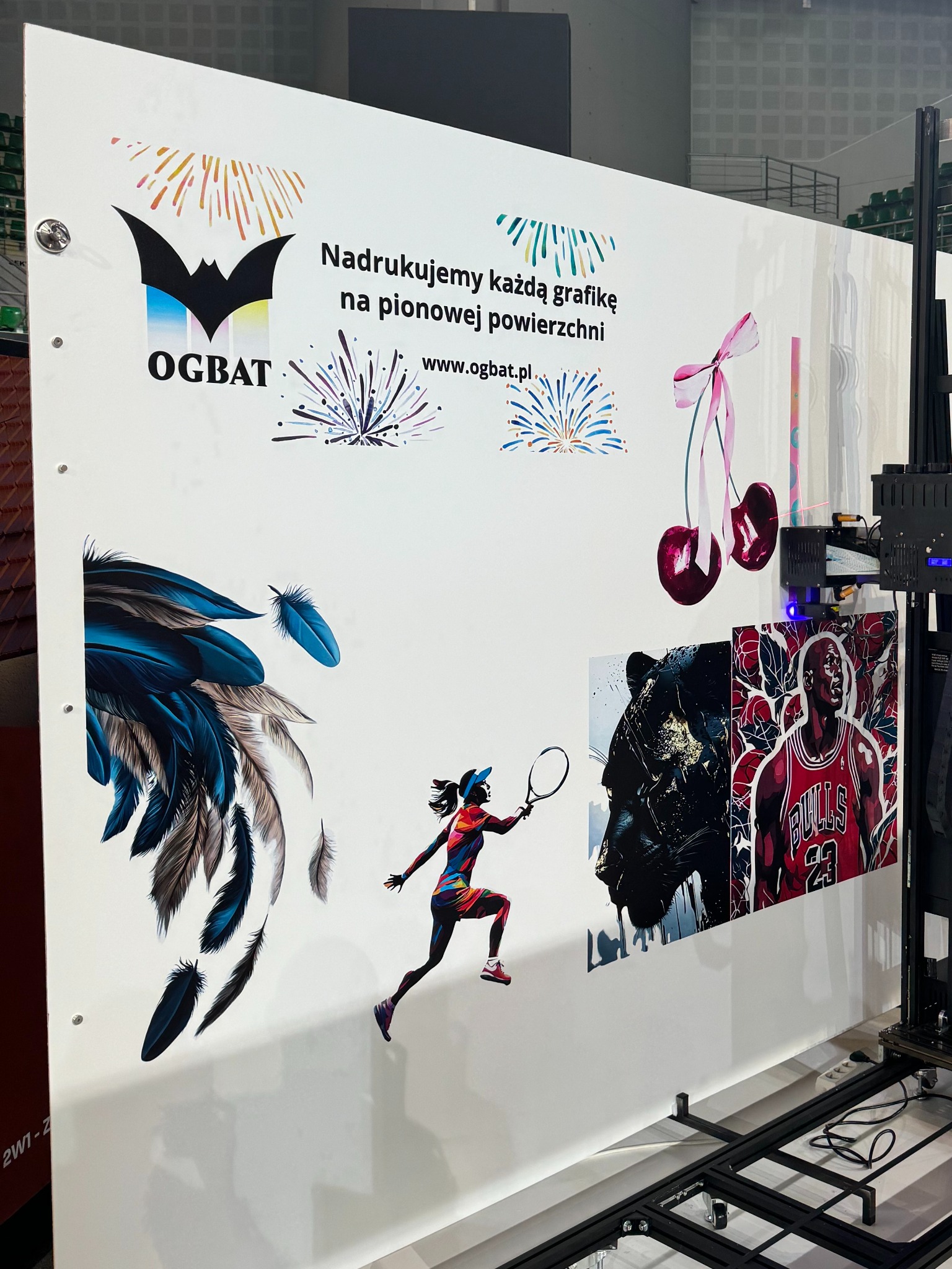 Wielkoformatowa drukarka pionowa nanosząca kolorowe grafiki na białą ścianę: logo firmy, fajerwerki, pióra, tenisistka, pantera i koszykarz Michael Jordan.