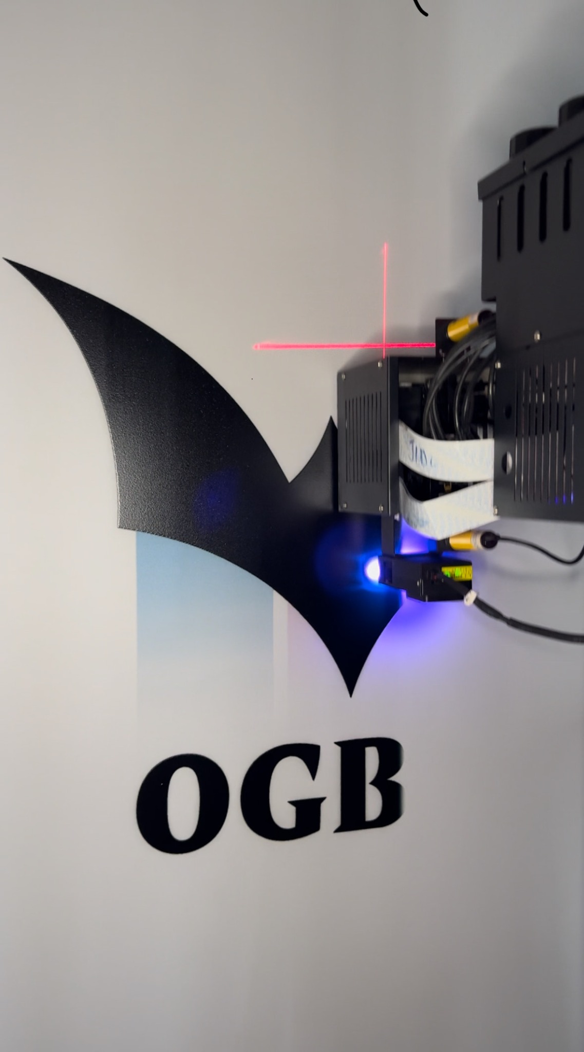 Aplikacja logo OGB na ścianie za pomocą projektora laserowego z niebieskim światłem i czerwoną linią celowniczą.