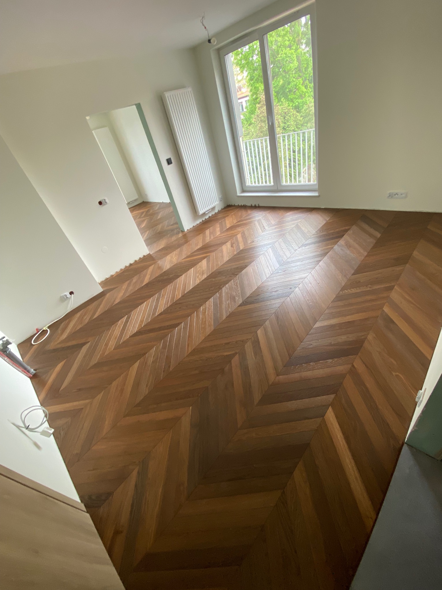 Ukończona podłoga z ciemnego, lakierowanego parkietu ułożonego we wzór jodełki francuskiej w jasnym pokoju z dużym oknem balkonowym i grzejnikiem.