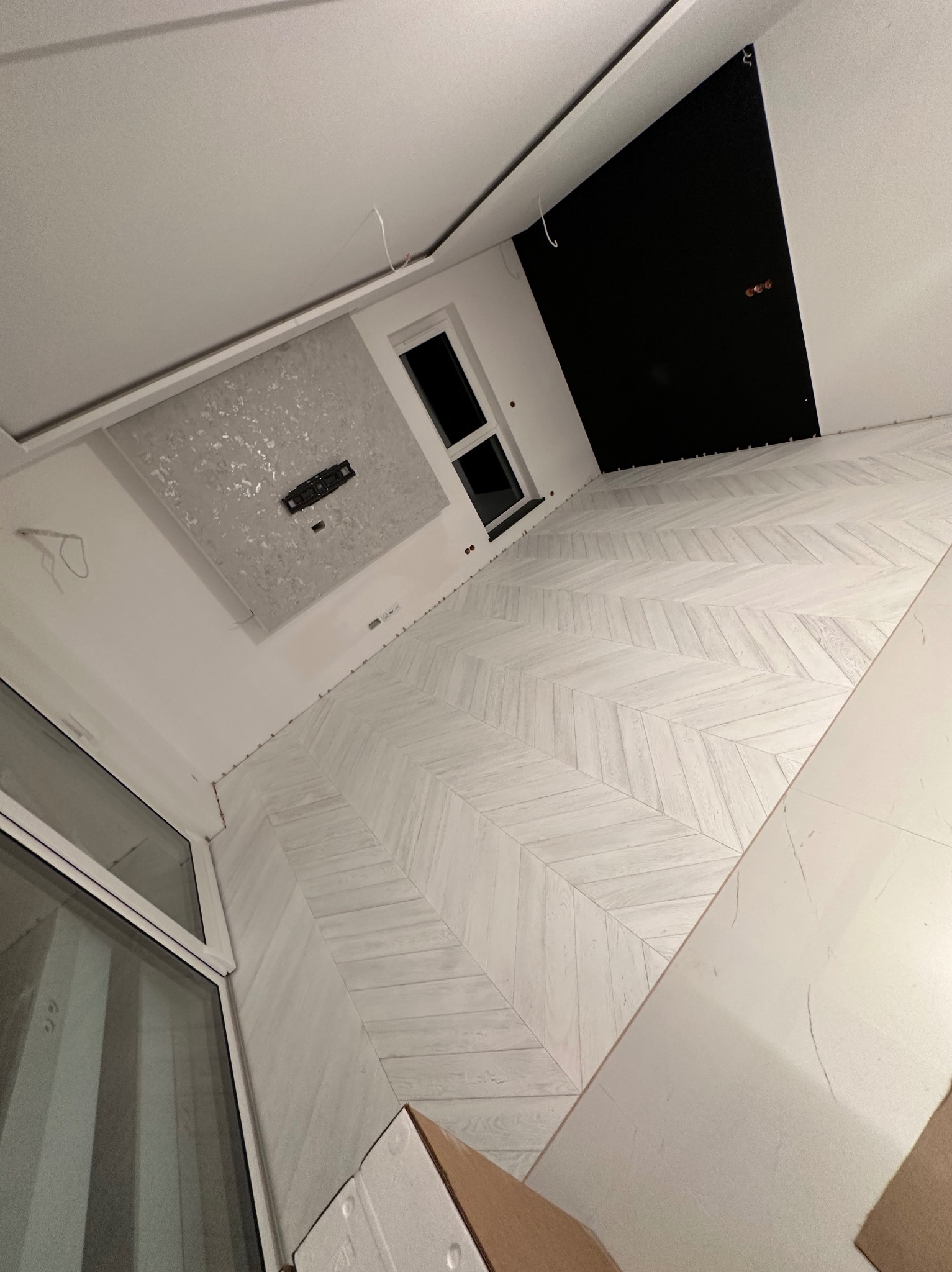 Wnętrze w trakcie wykańczania: jasne panele ułożone w jodełkę francuską, widoczne fragmenty okna, sufitu z elementami dekoracyjnymi i czarną ścianą, brak listew przypodłogowych.