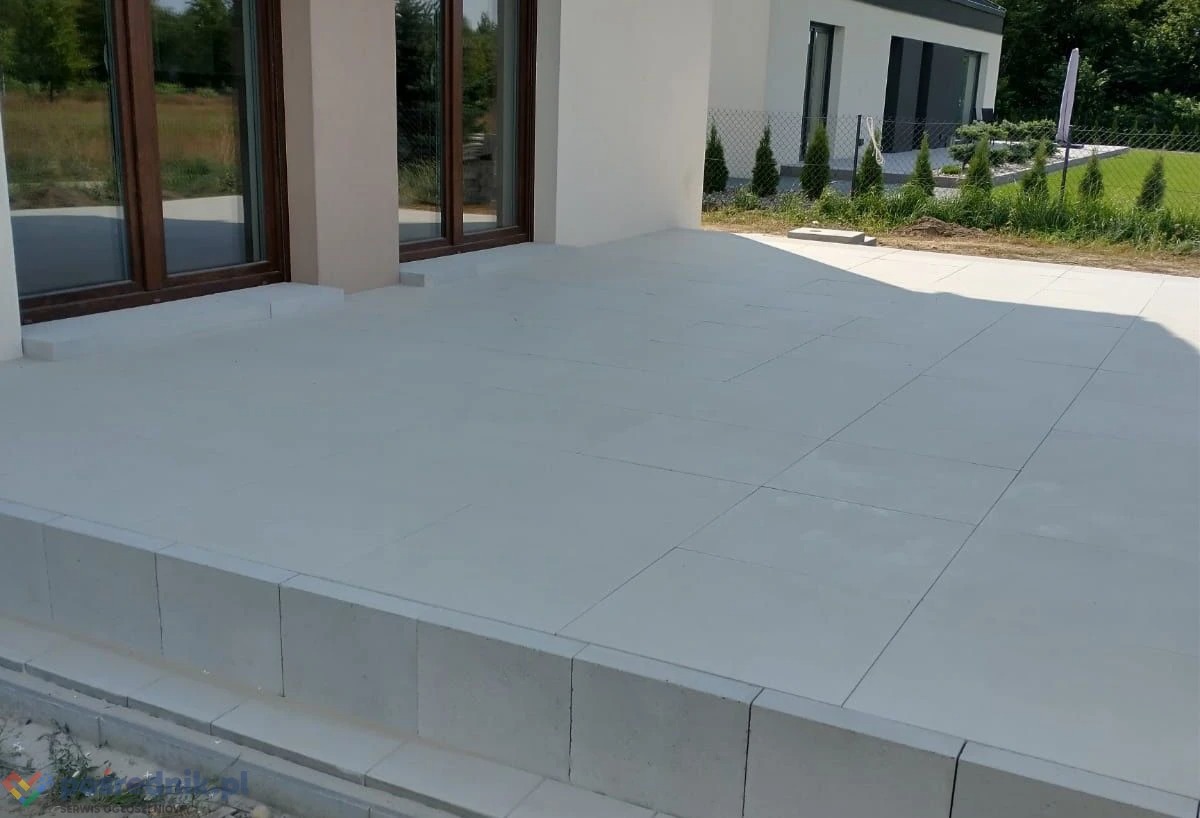 Jasnoszara kostka brukowa ułożona na tarasie przed nowoczesnym domem z dużymi oknami, widoczne schody z kostki brukowej i fragment ogrodu z tujami.