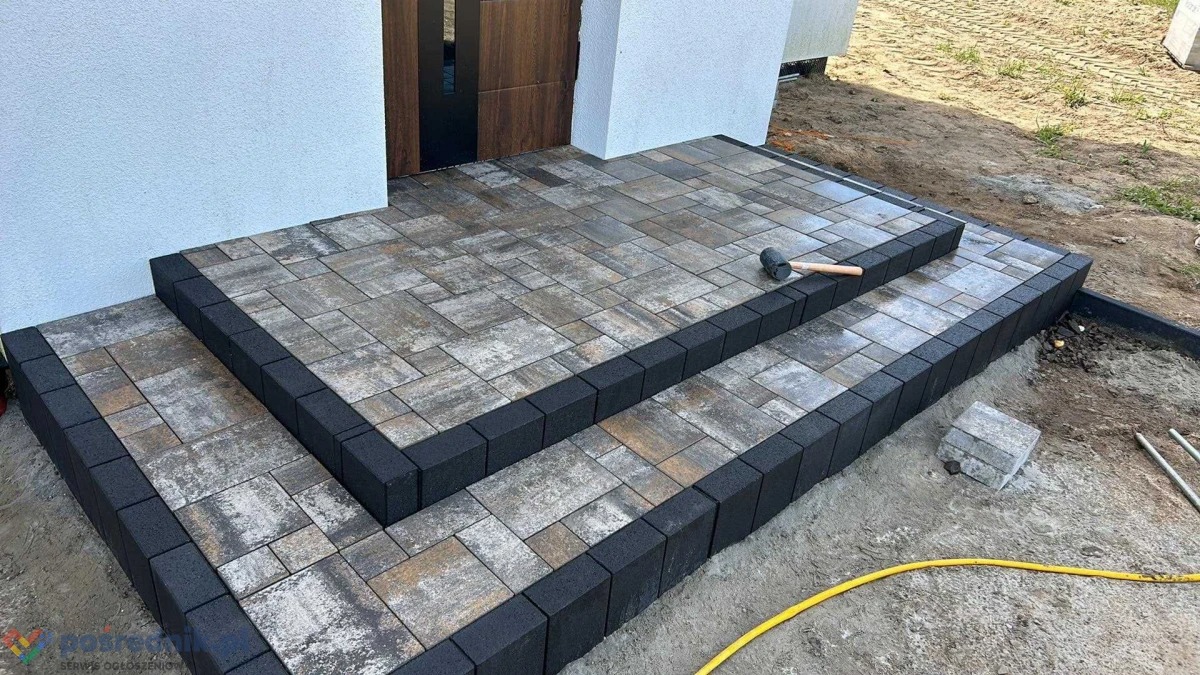 Schody wejściowe do domu wyłożone kostką brukową w odcieniach szarości i brązu, obrzeża z ciemnoszarej kostki, widoczny gumowy młotek i fragment żółtego węża ogrodowego.