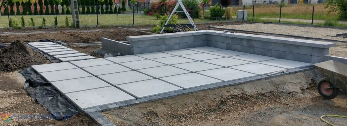 Układanie kwadratowych płyt chodnikowych na przygotowanym podłożu, widoczny fragment murku oporowego z szarej kostki, w tle ogród i dom w budowie.